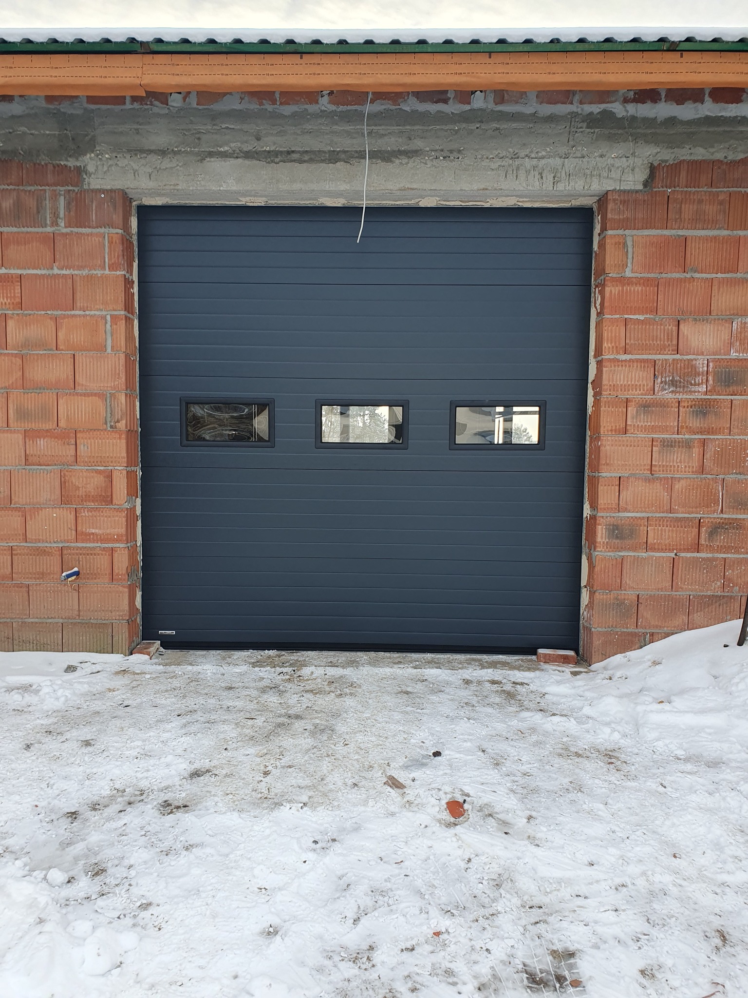 Grafitowa brama garażowa segmentowa z trzema okienkami, zamontowana w surowym murze z czerwonej cegły. Przed bramą leży śnieg.