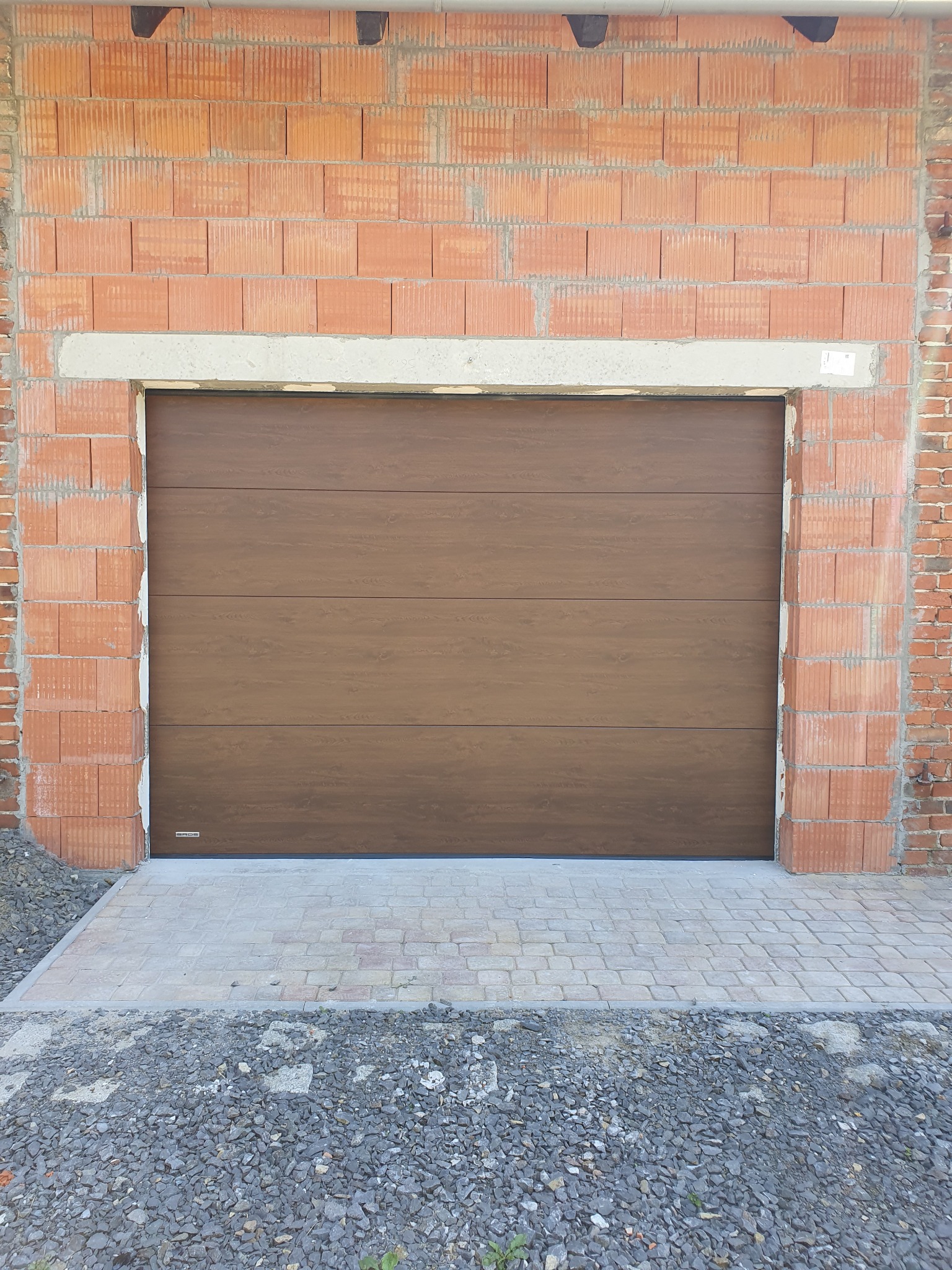 Brązowa brama garażowa segmentowa z poziomymi przetłoczeniami, zamontowana w surowym murze z cegły dziurawki. Widoczny fragment podjazdu z kostki i kamienia.