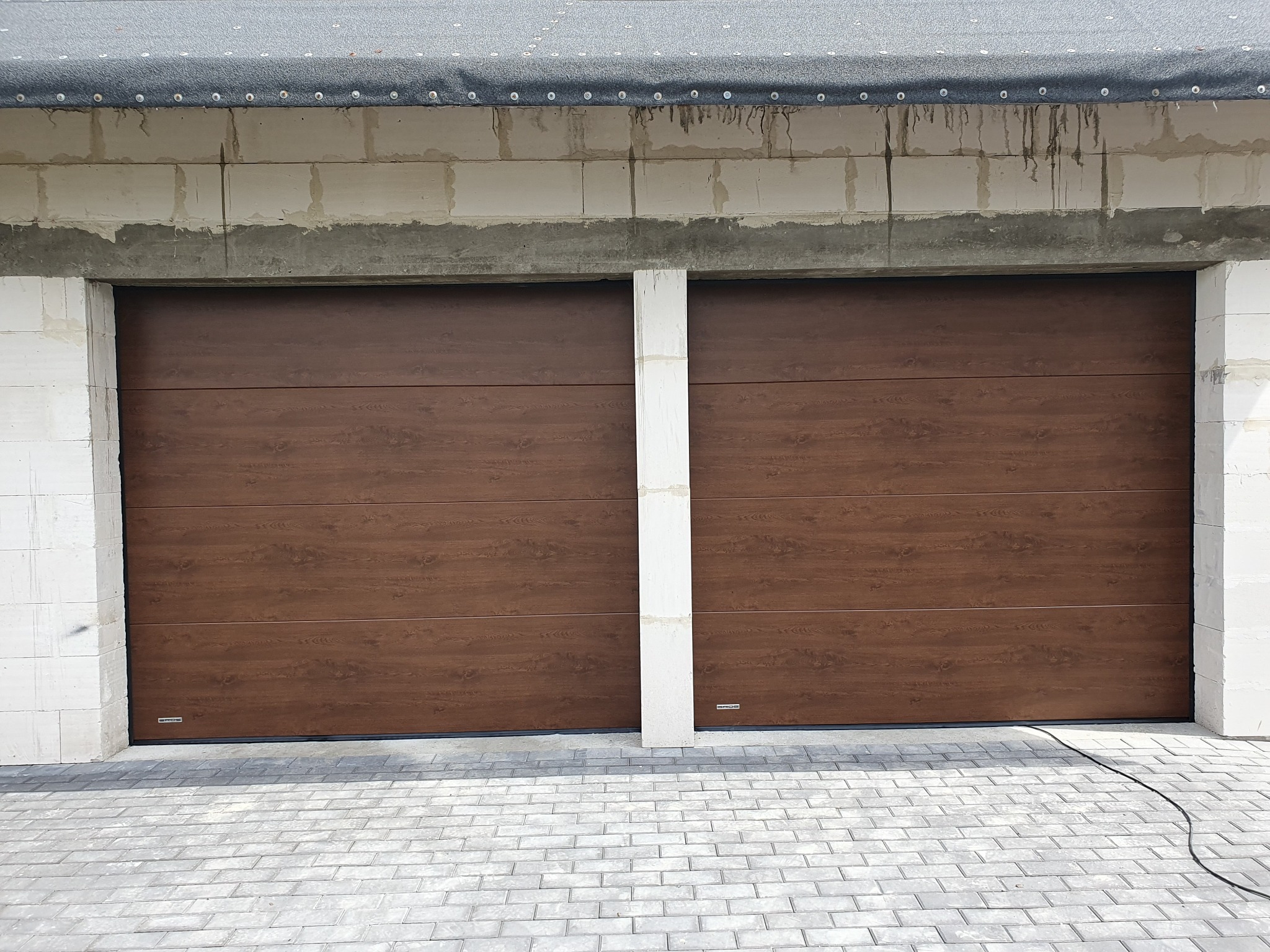 Dwie brązowe bramy garażowe segmentowe z widoczną fakturą drewna, zamontowane w surowym budynku z bloczków betonowych, na tle szarej kostki brukowej.