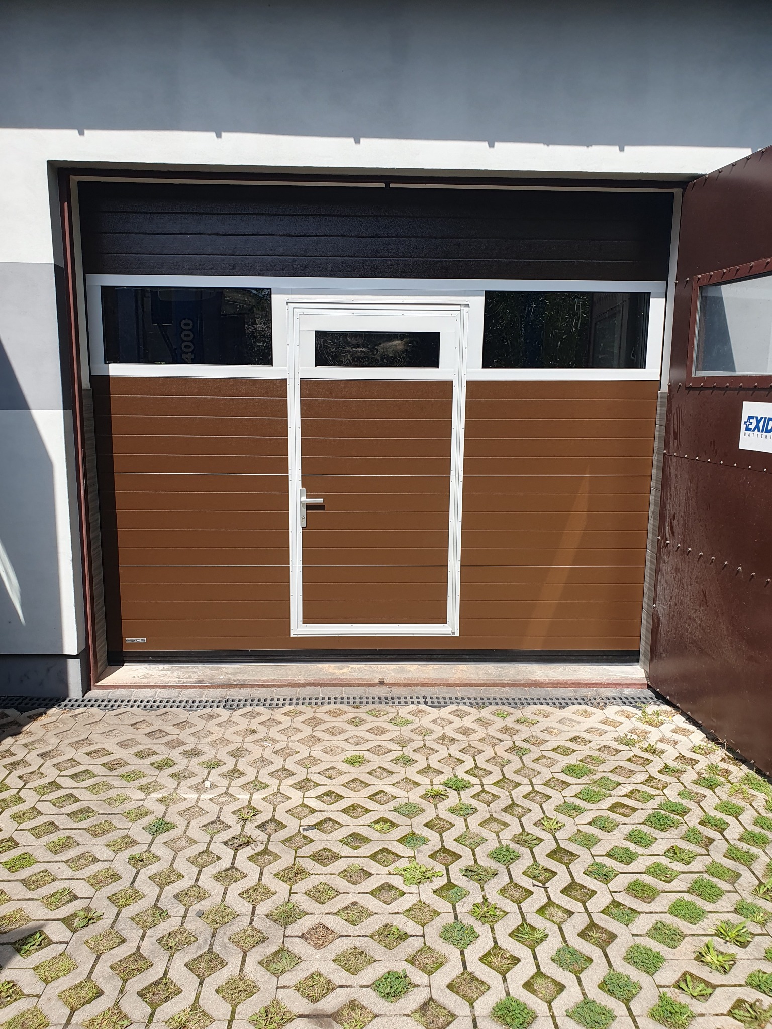 Segmentowa brama garażowa w odcieniach brązu z oknami i drzwiami wejściowymi, częściowo otwarta, z widoczną kostką brukową na podjeździe.