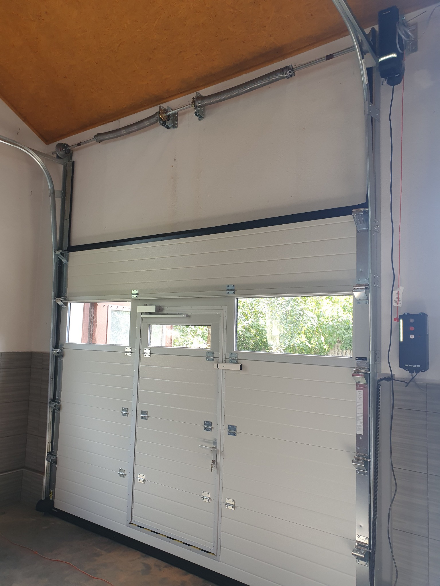 Otwarta segmentowa brama garażowa z drzwiami i oknami, widok od wewnątrz garażu, mechanizm sprężynowy i napęd elektryczny widoczne.