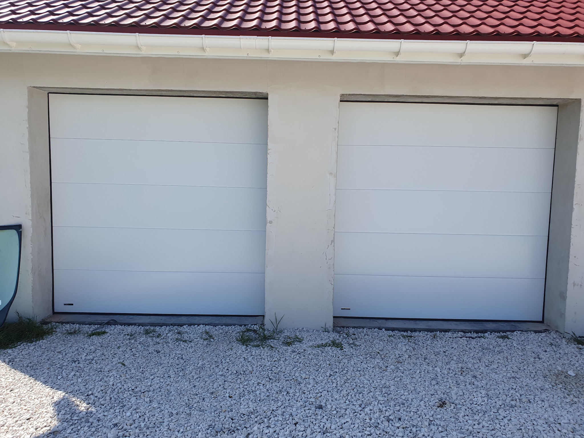 Dwie białe, segmentowe bramy garażowe w jasnym garażu z czerwonym dachem, otoczone białym żwirem. Minimalistyczny design, symetryczny układ.
