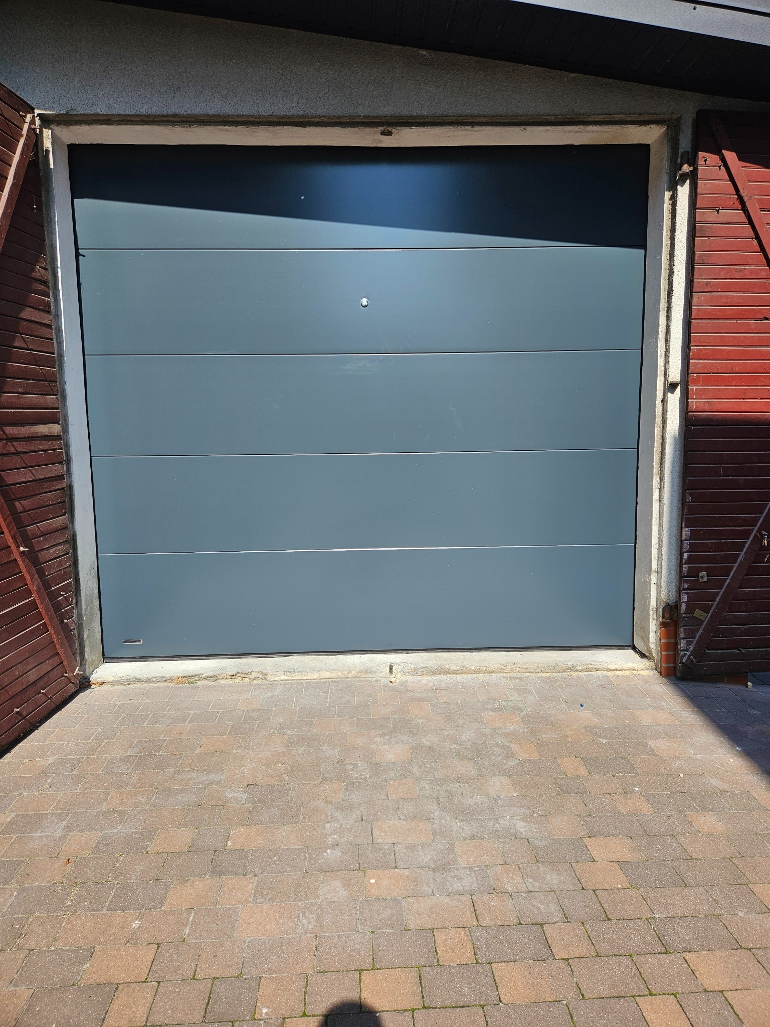 Szara, segmentowa brama garażowa z poziomymi panelami, widoczna z zewnątrz na tle kostki brukowej i fragmentów drewnianej konstrukcji garażu.