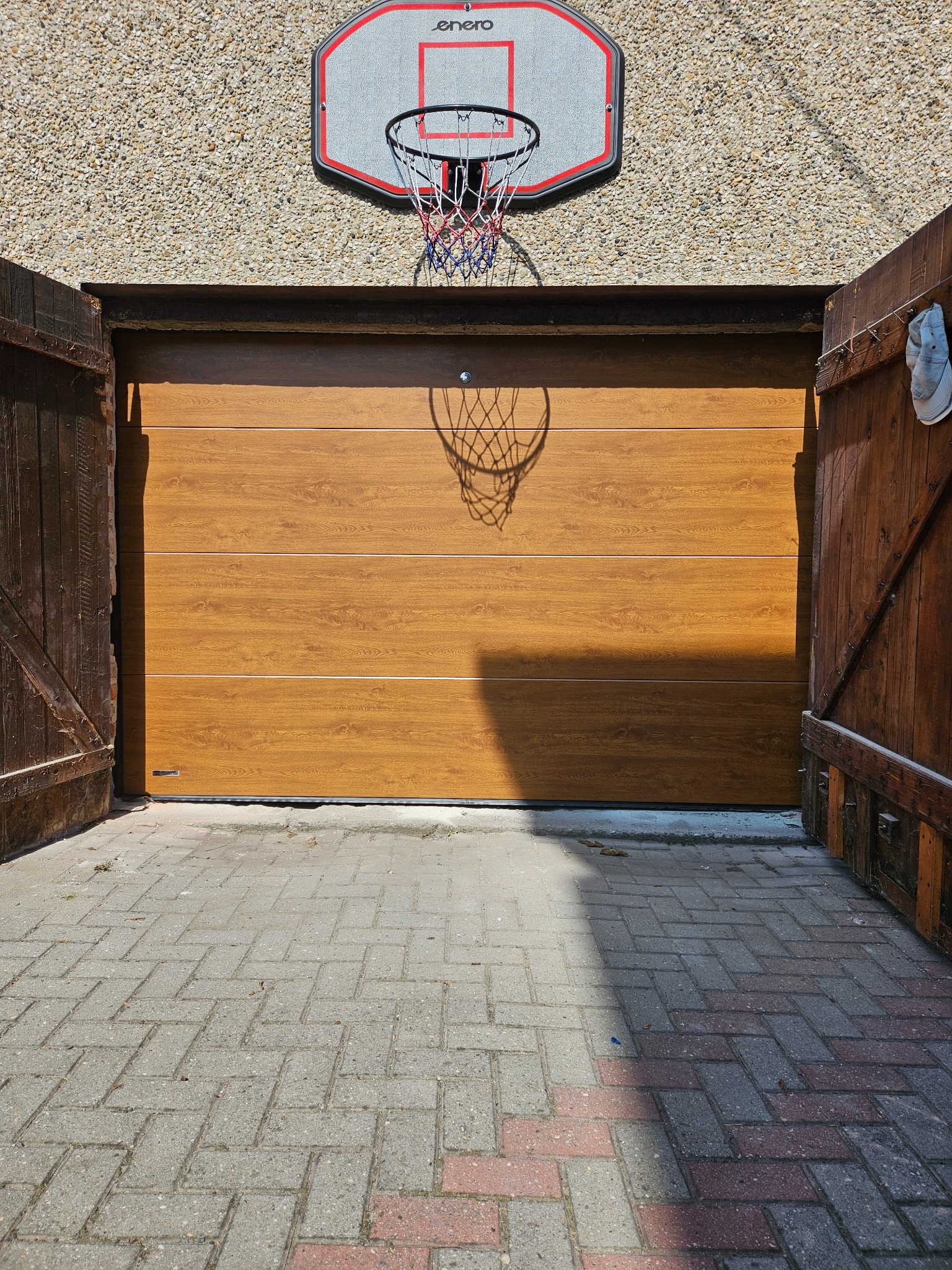 Otwarta, segmentowa brama garażowa w odcieniach brązu, z koszem do koszykówki nad nią. Widoczne drewniane, rozwarte wrota i kostka brukowa przed wjazdem.
