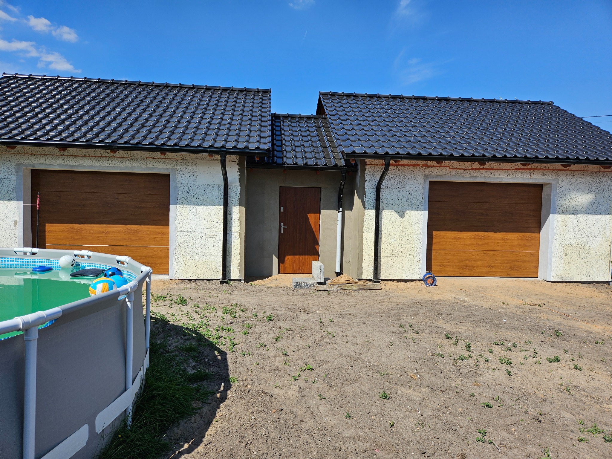 Nowy budynek gospodarczy z dwiema drewnopodobnymi bramami garażowymi i drzwiami wejściowymi, na pierwszym planie częściowo widoczny basen ogrodowy.