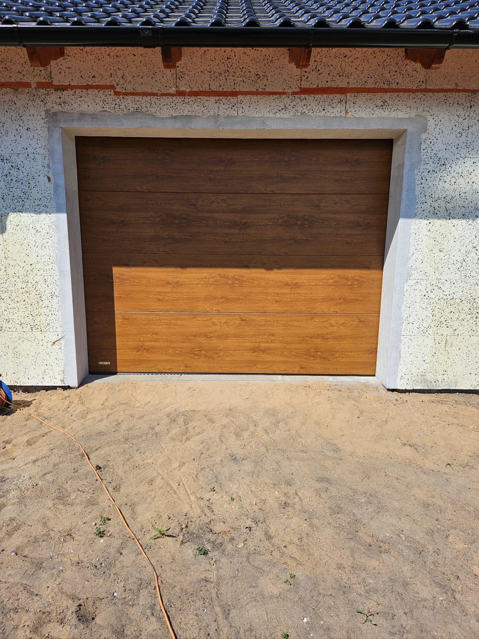 Zamknięta, segmentowa brama garażowa w kolorze drewna, zamontowana w surowym otworze budynku. Widoczne detale wykończenia i faktura bramy.