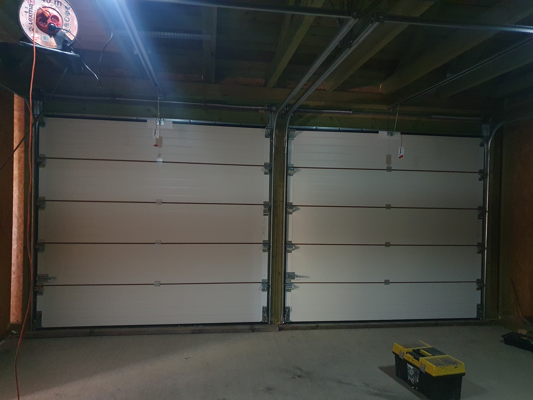Zamknięta, segmentowa brama garażowa wewnątrz garażu z widoczną skrzynką narzędziową. Widoczne elementy montażowe i konstrukcja bramy.