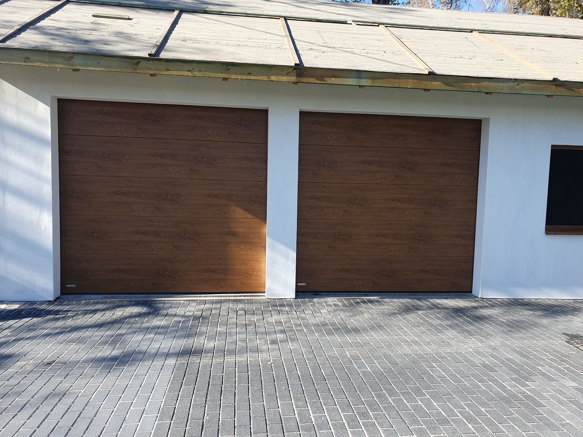 Dwie brązowe bramy garażowe segmentowe z poziomym wzorem drewna, zamontowane w białym budynku z szarą kostką brukową na podjeździe. Widoczne detale wykończenia.
