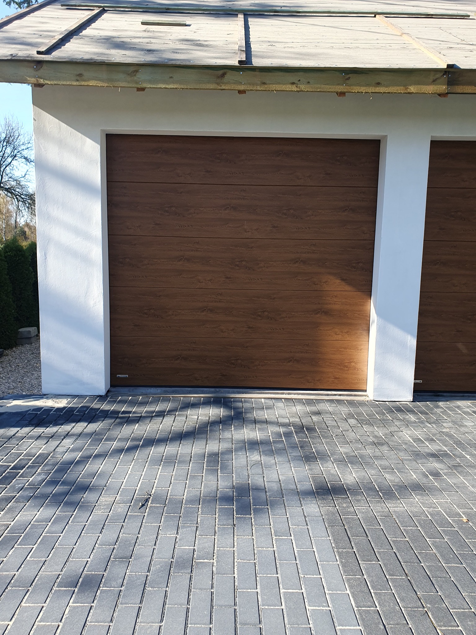 Nowoczesna brama garażowa segmentowa w kolorze drewna, z widoczną fakturą usłojenia, zamontowana w białej elewacji budynku. Kostka brukowa przed bramą.