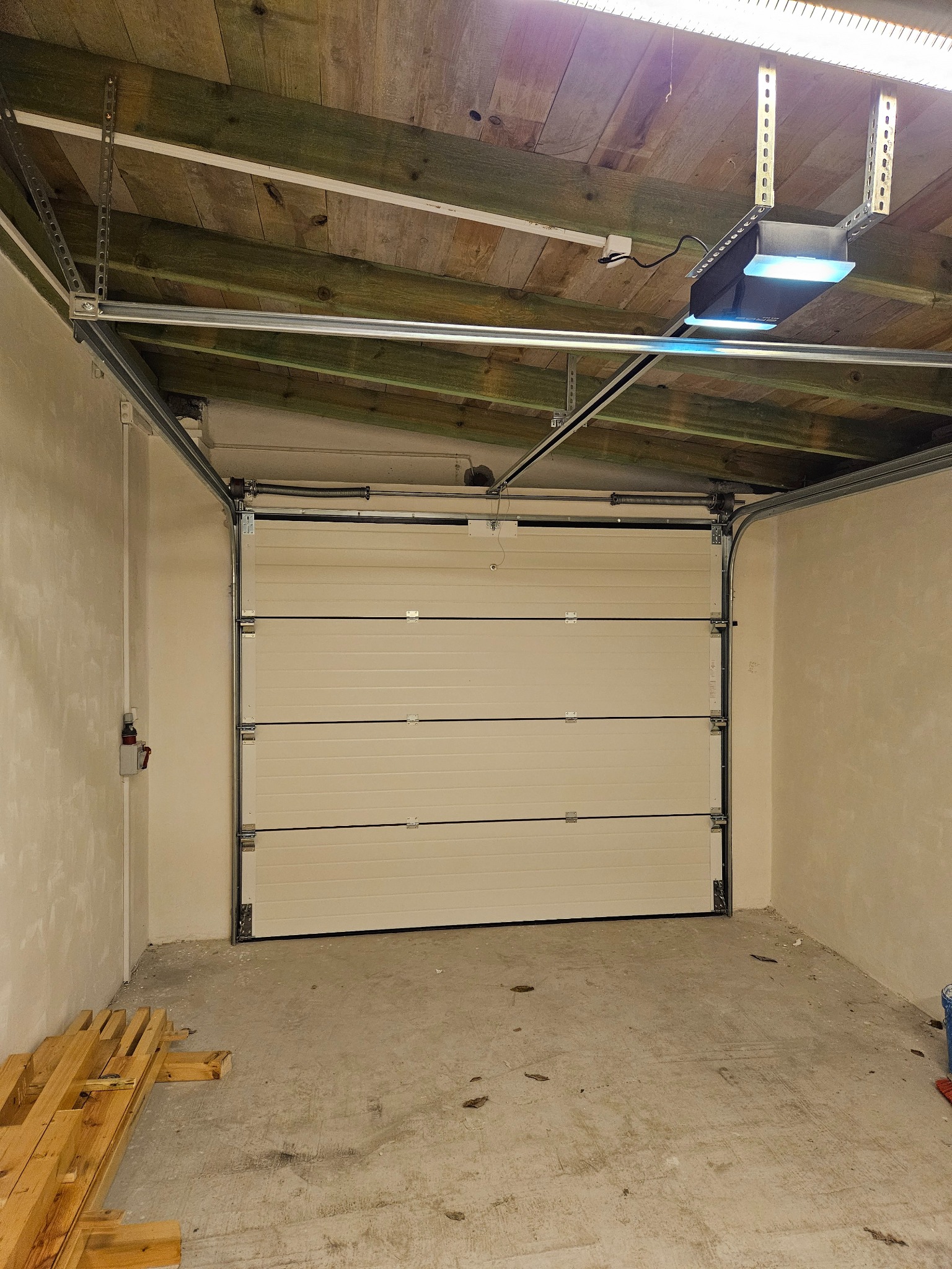 Zamknięta, segmentowa brama garażowa w jasnym odcieniu, widoczny mechanizm otwierania pod drewnianym sufitem garażu, surowy beton na podłodze.