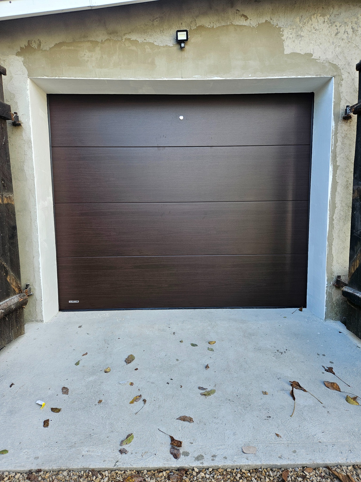 Ciemnobrązowa, segmentowa brama garażowa z poziomymi panelami, zamontowana w surowym otworze garażowym z widocznymi śladami prac murarskich i betonową posadzką.