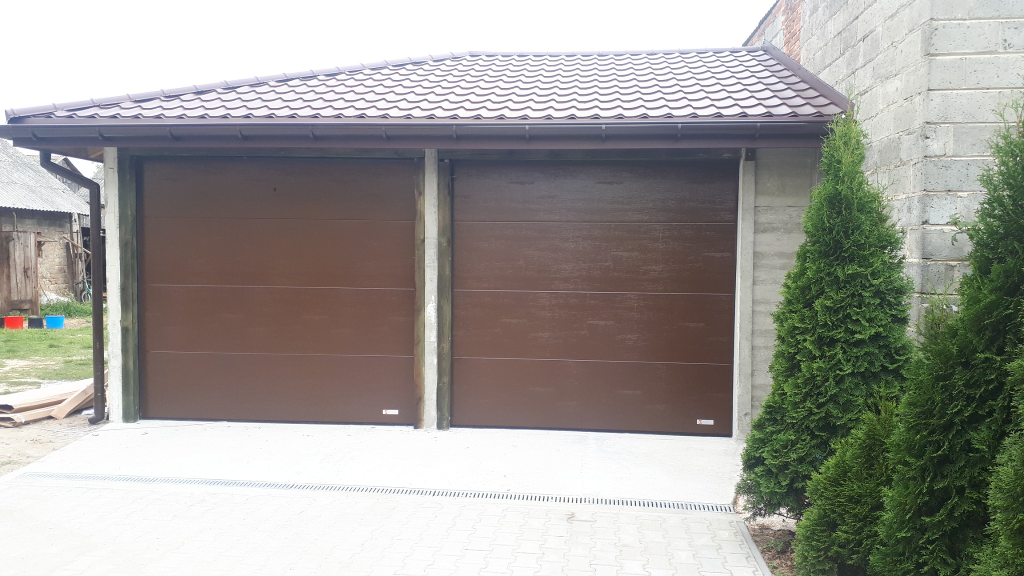 Dwie brązowe, segmentowe bramy garażowe zadaszone brązowym dachem, widoczne z zewnątrz, obok rosną tuje.