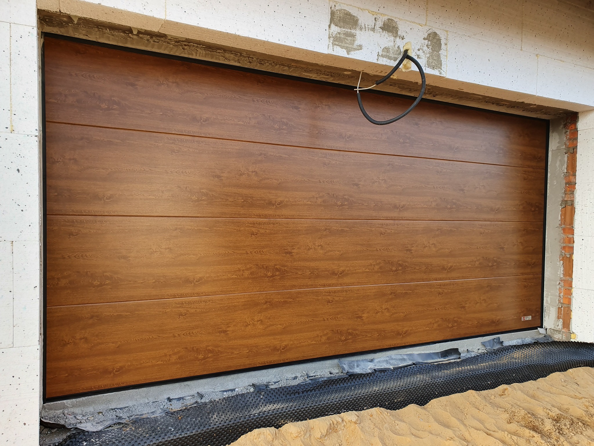 Zamknięta segmentowa brama garażowa w kolorze drewna, widoczna na tle surowych ścian budynku w trakcie budowy.