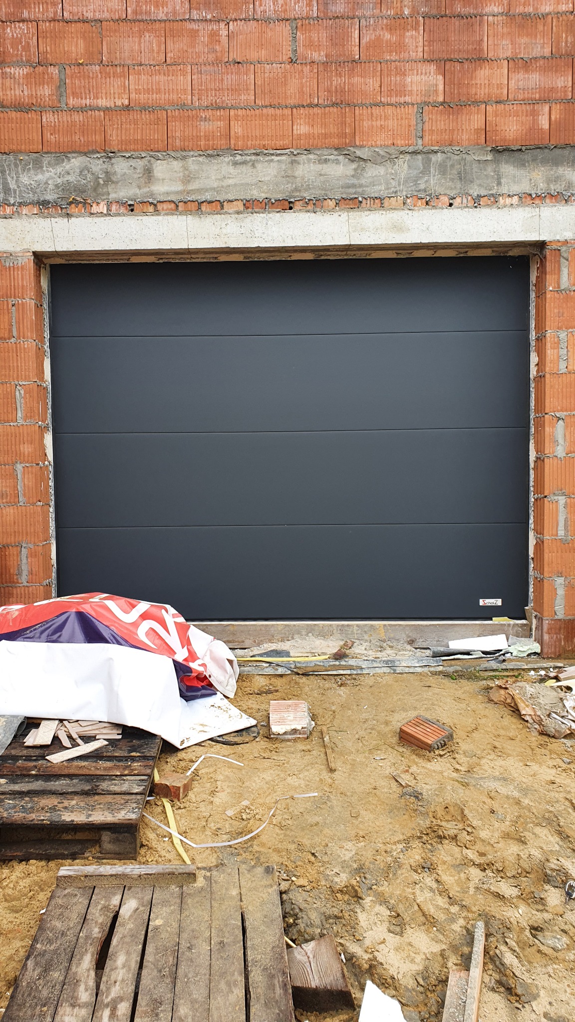 Szara segmentowa brama garażowa zamontowana w surowym otworze budynku z czerwonej cegły, widoczne palety i materiały budowlane na ziemi.