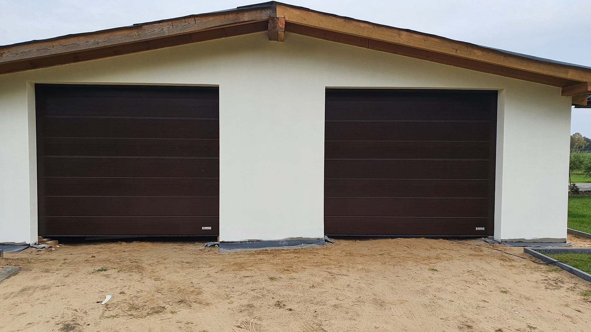 Dwie brązowe segmentowe bramy garażowe w jasnym budynku z drewnianą konstrukcją dachu, widoczne niedokończone prace brukarskie przed wjazdem.