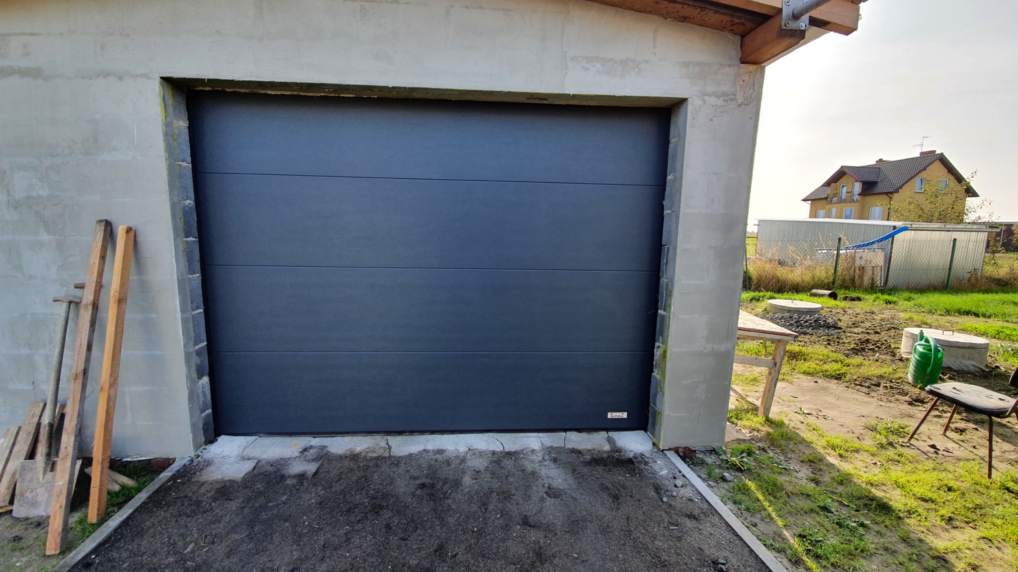 Szara segmentowa brama garażowa w surowym otoczeniu, z widocznymi narzędziami budowlanymi i niedokończonymi ścianami garażu.