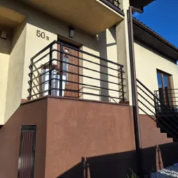 Nowoczesna, czarna balustrada balkonowa z poziomymi szczeblami, zamontowana przy budynku z numerem 50B. Widoczne kwiaty w donicach na balkonie.