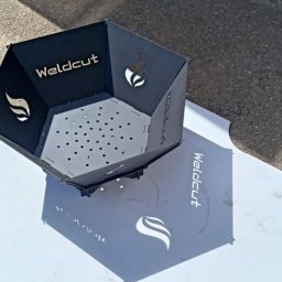 Weldcut spawanie i cięcie s.c. - Metalowy kosz na ognisko z wyciętym laserowo logo i wzorem płomienia, ustawiony na białym arkuszu na zewnątrz. Widoczne perforowane dno i cień.