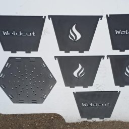 Weldcut spawanie i cięcie s.c. - Części wycięte z metalu na palniku CNC, gotowe do spawania. Elementy z logo i motywem płomienia, leżą na białym tle. Widoczny fragment podłoża.