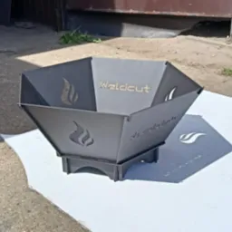 Metalowy kosz na ognisko z wyciętym logo 'Weldcut' i motywem płomieni, ustawiony na białej powierzchni na zewnątrz.