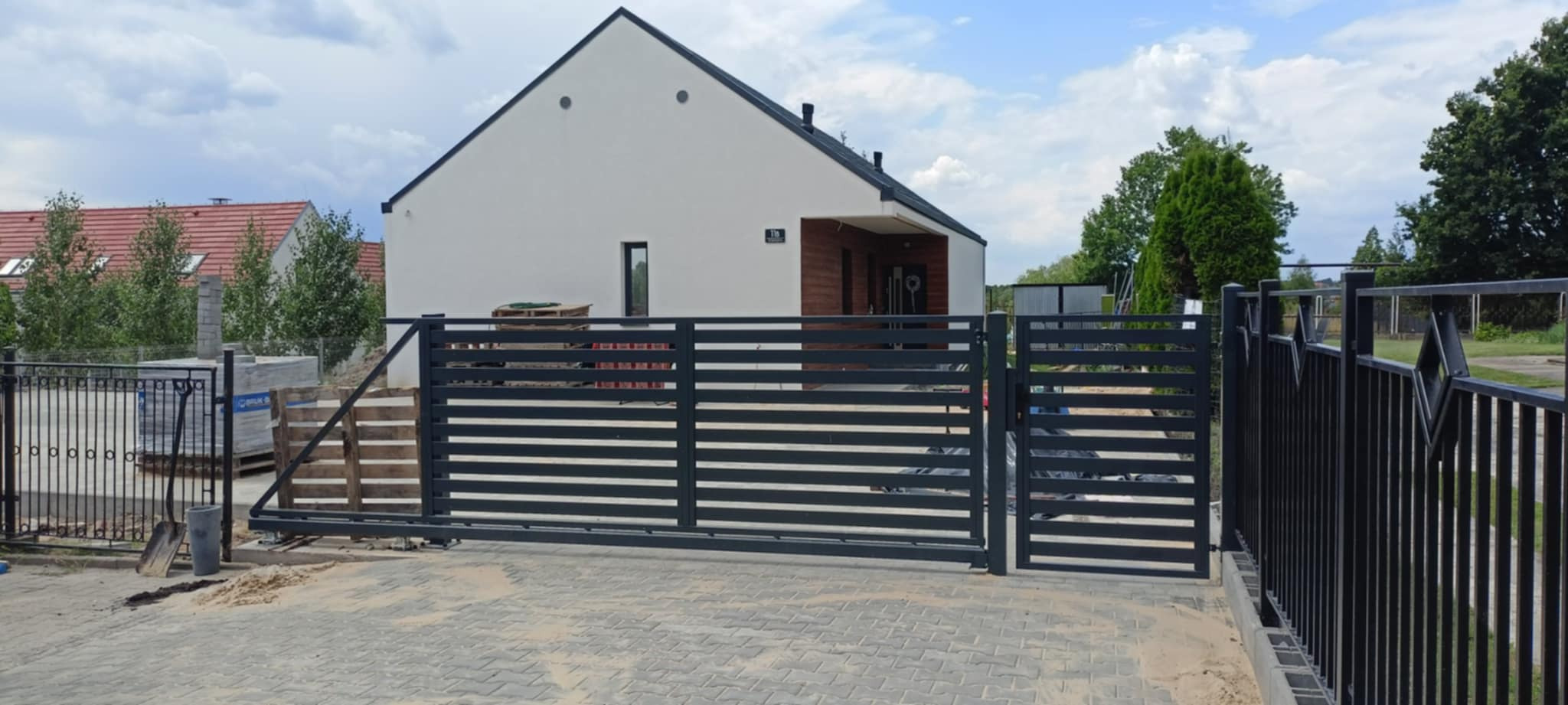 Nowoczesna, przesuwna brama w kolorze grafitowym z poziomymi panelami, na tle jasnego domu jednorodzinnego i kostki brukowej. Widoczny fragment ogrodzenia.