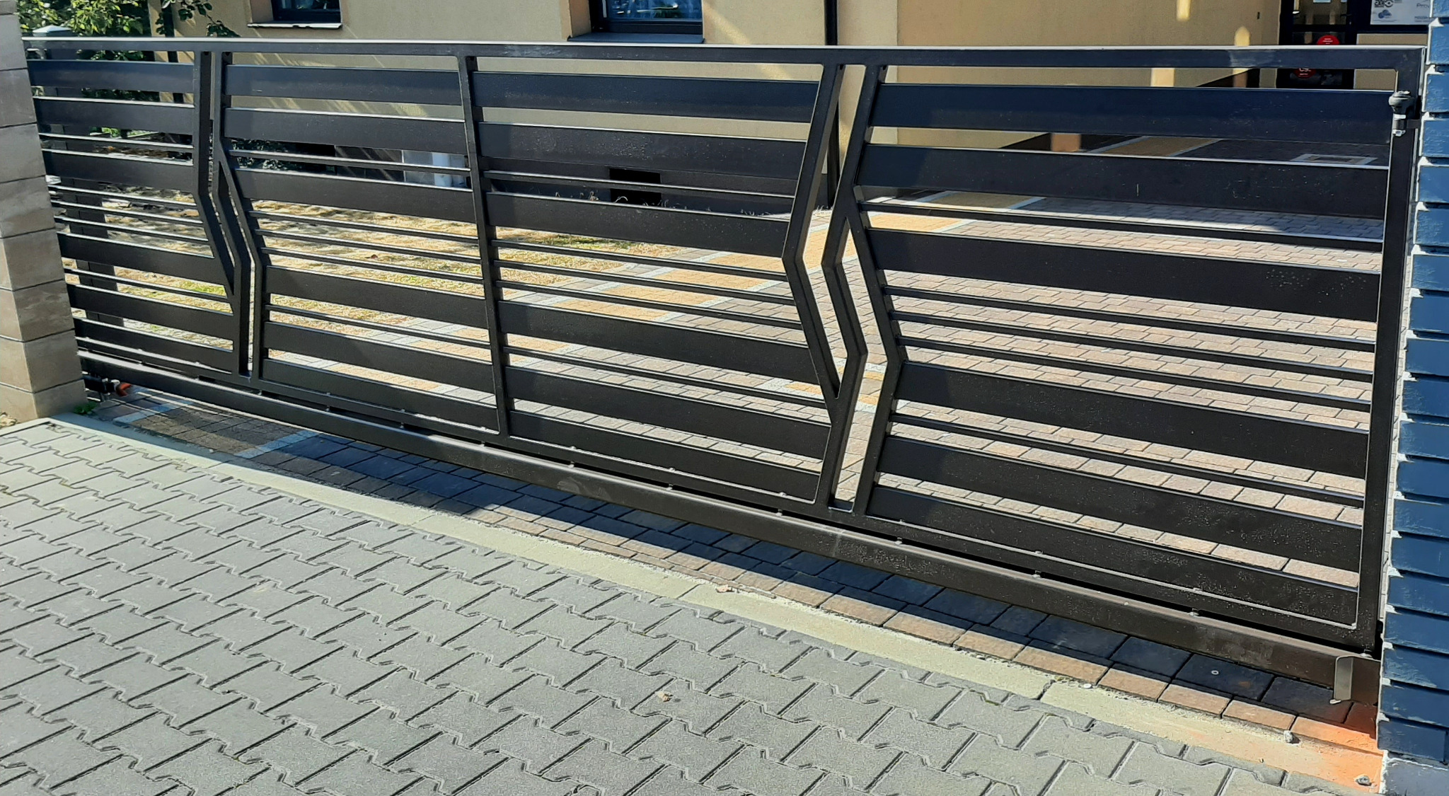 Nowoczesna, przesuwna brama w kolorze antracytu z poziomymi profilami i dekoracyjnymi elementami, zamontowana na posesji z kostką brukową.