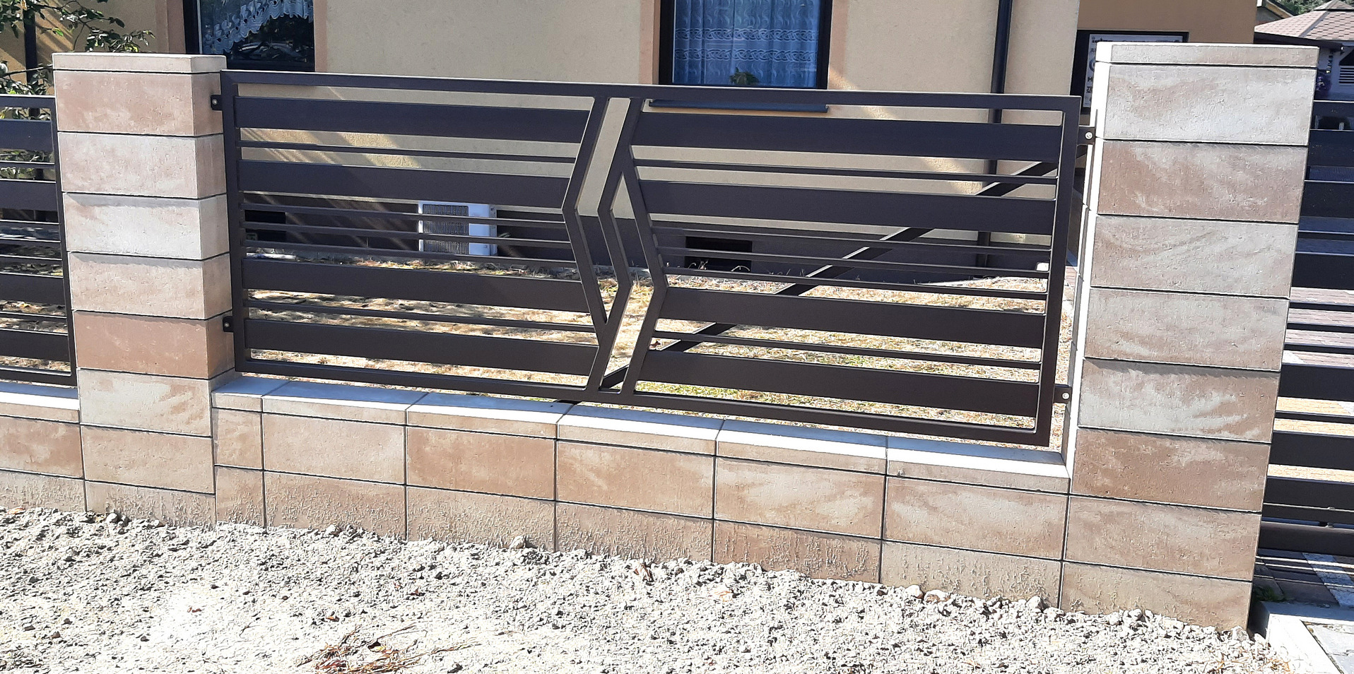 Nowoczesne ogrodzenie metalowe z poziomymi przęsłami i geometrycznym wzorem, osadzone na niskim murku z betonowych bloczków.