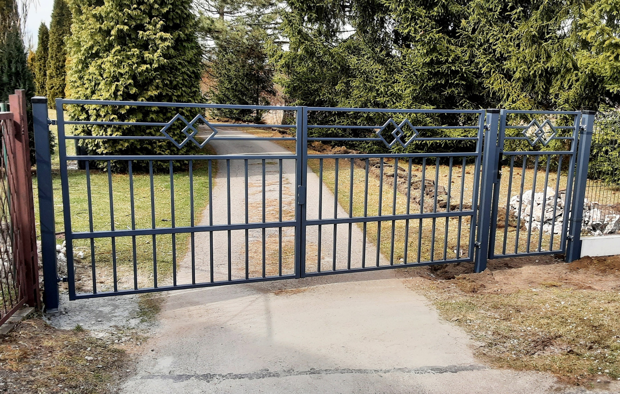 Metalowa brama wjazdowa w kolorze grafitowym z geometrycznym wzorem rombów, prowadząca do posesji z zielenią w tle. Widok na ścieżkę.
