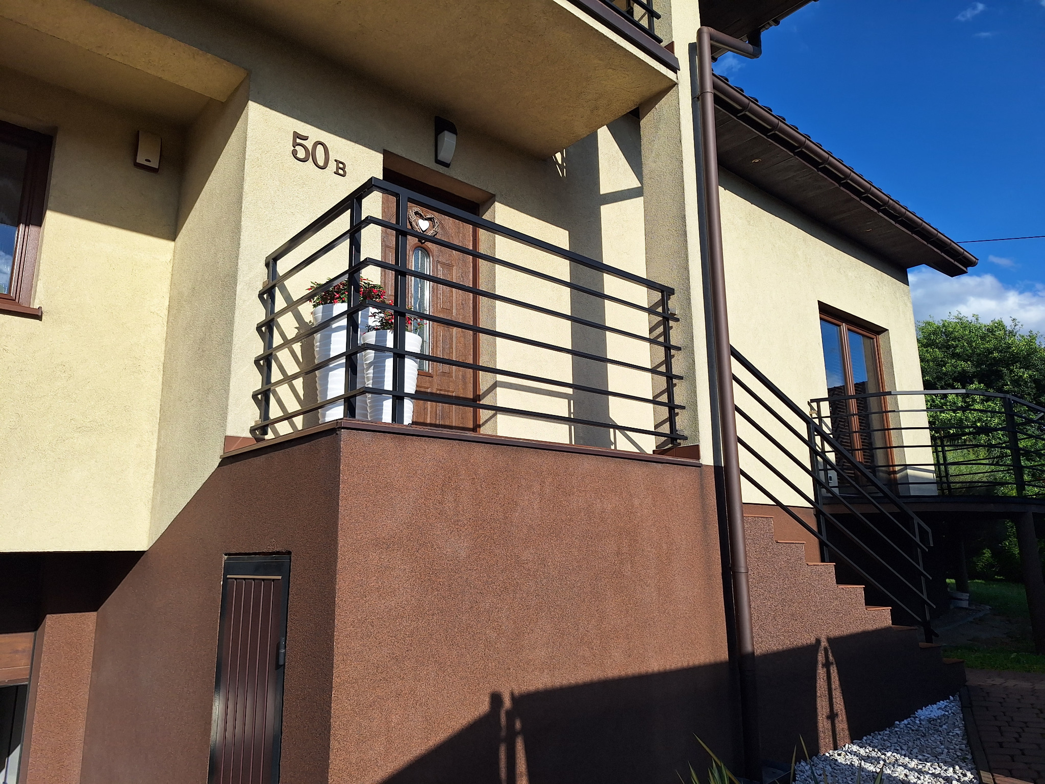 Nowoczesna, czarna balustrada balkonowa z poziomymi szczeblami, zamontowana przy budynku z numerem 50B. Widoczne kwiaty w donicach na balkonie.