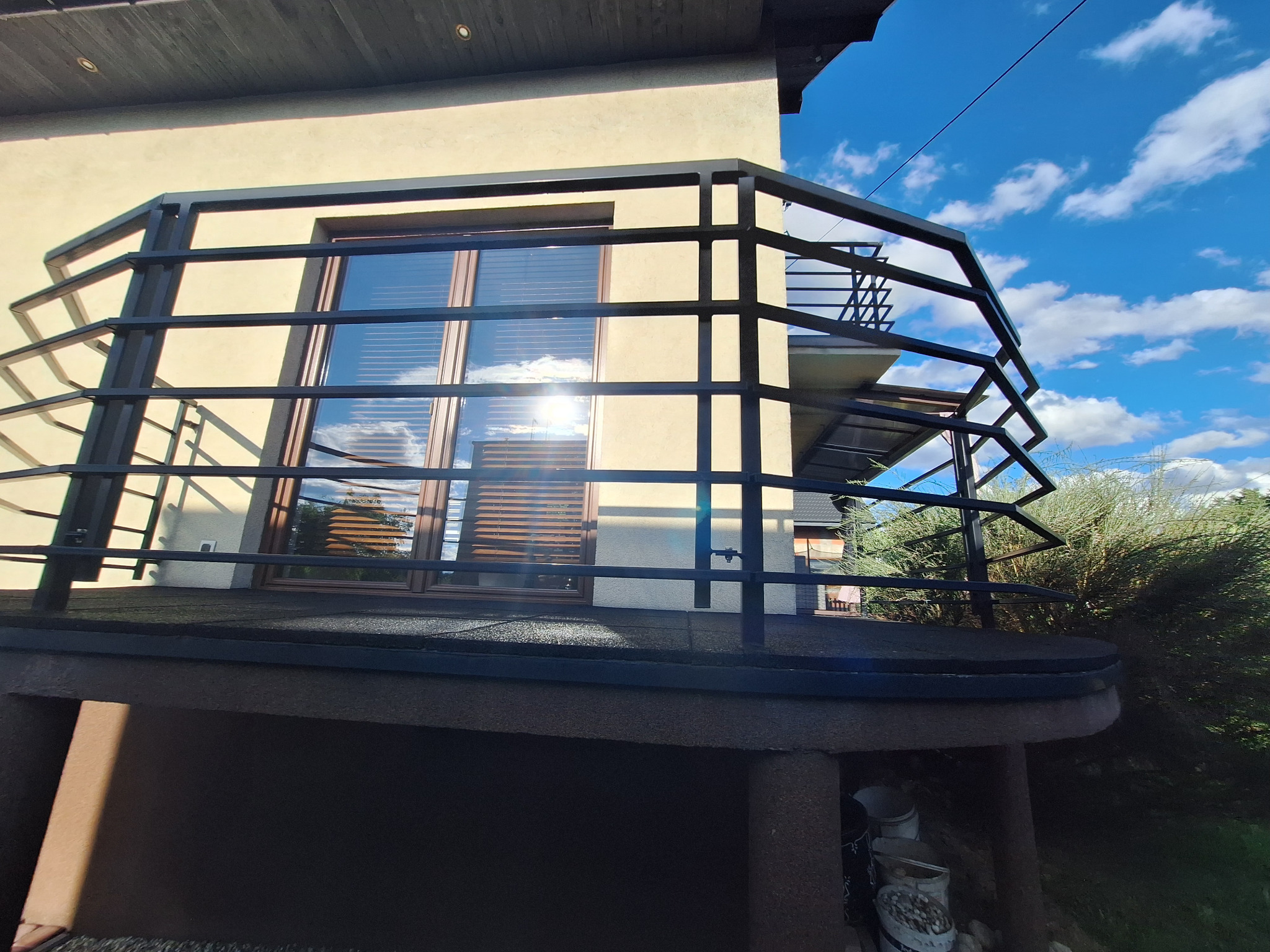 Nowoczesna, czarna balustrada balkonowa z metalu, widok z dołu, z refleksami słońca na oknach. Minimalistyczny design i błękitne niebo w tle.