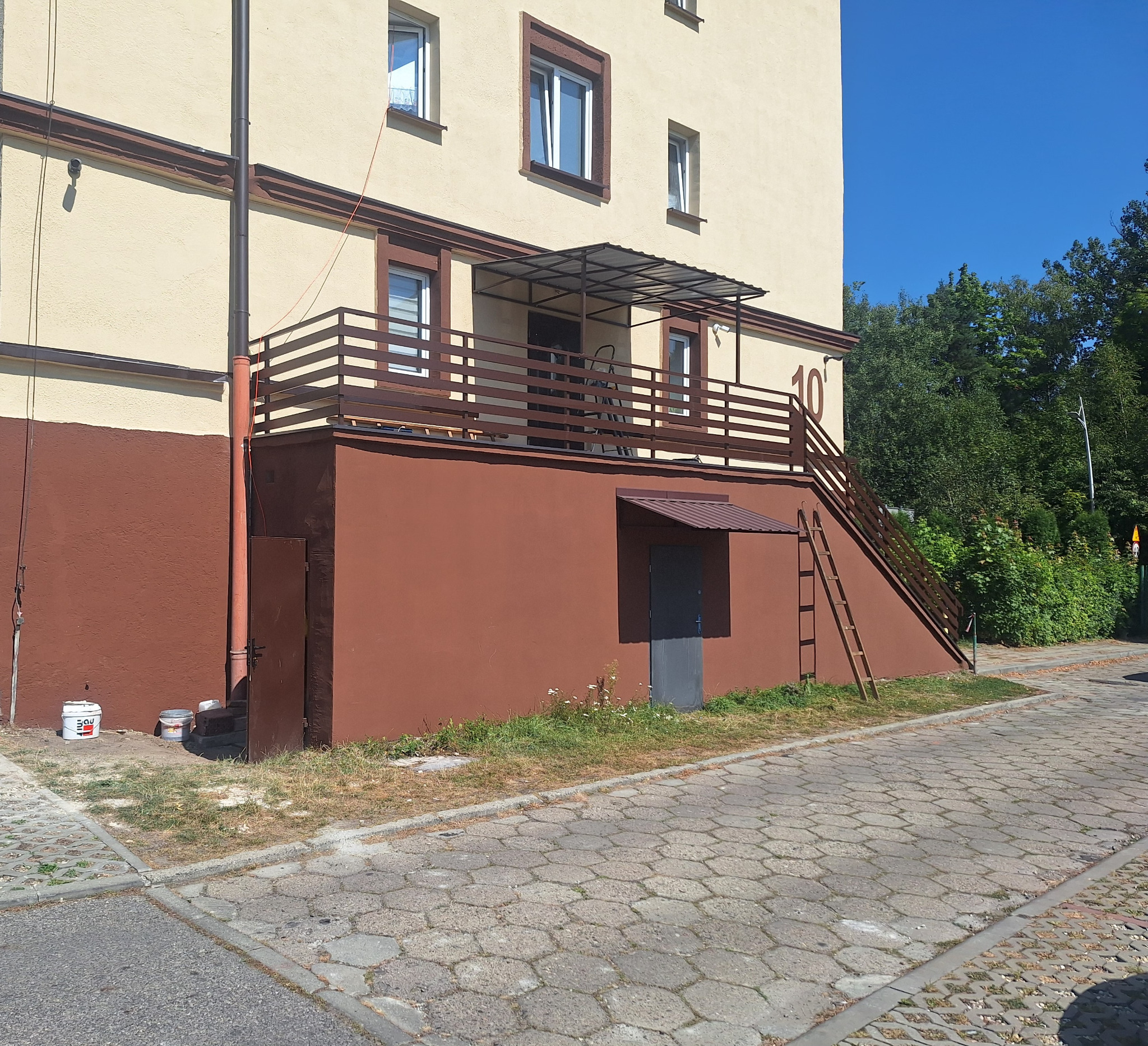 Balkon z drewnianą balustradą i schodami na zewnątrz budynku. Widoczna drabina oparta o schody i drzwi do piwnicy z daszkiem. Jasne tło budynku.