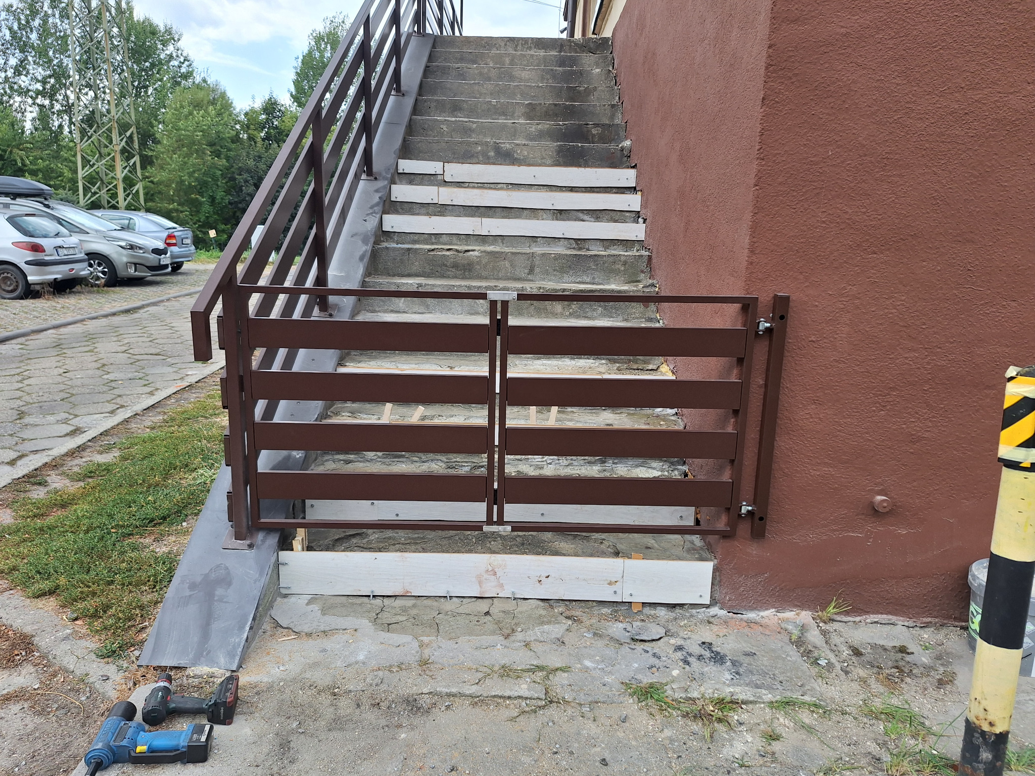 Montaż brązowej, metalowej furtki schodowej z poziomymi szczebelkami. W tle schody betonowe z brązową balustradą i narzędzia budowlane.