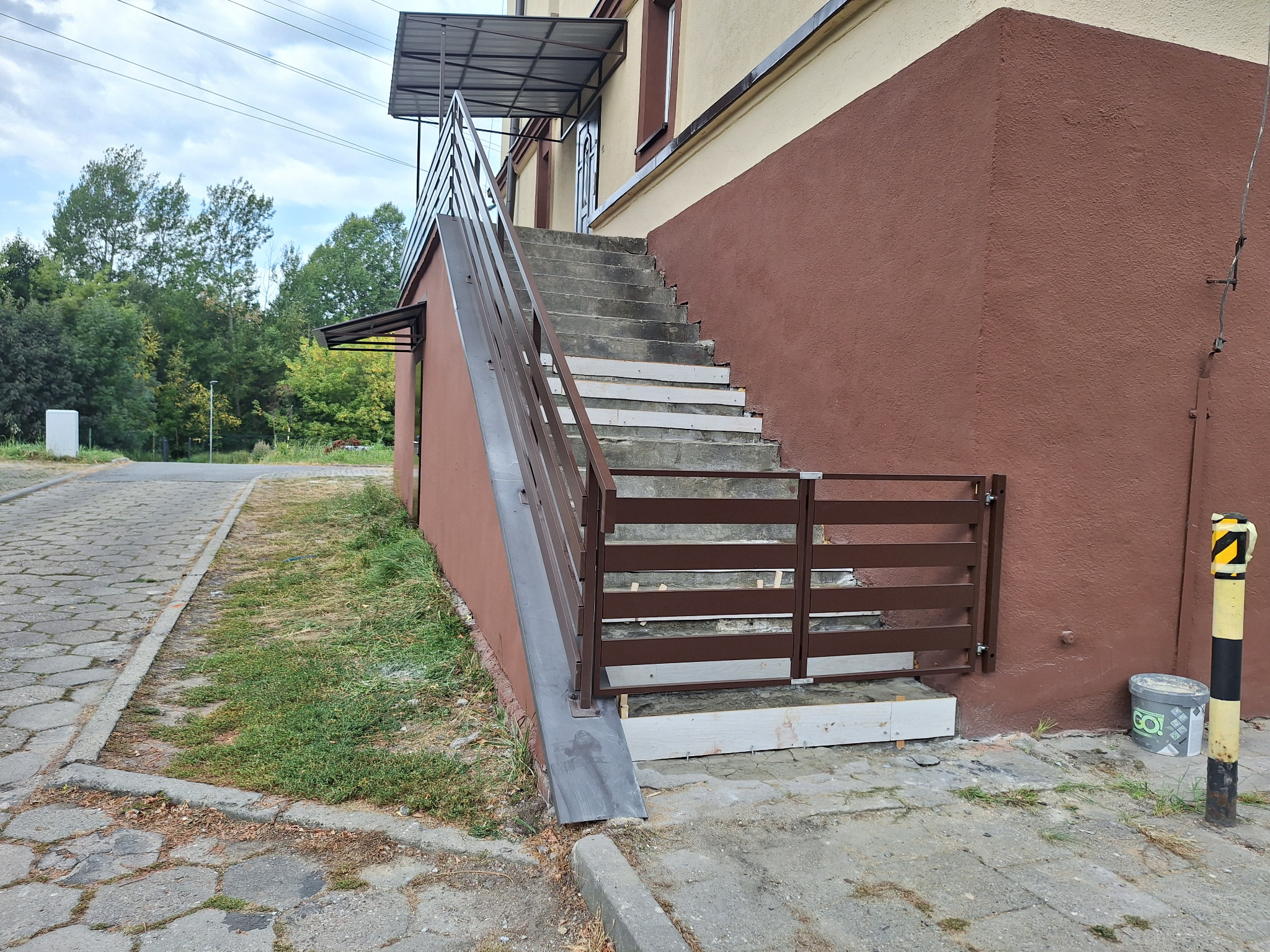 Zewnętrzne schody betonowe z brązową metalową poręczą i barierką, zadaszone. Widoczne elementy wykończeniowe i otoczenie budynku.