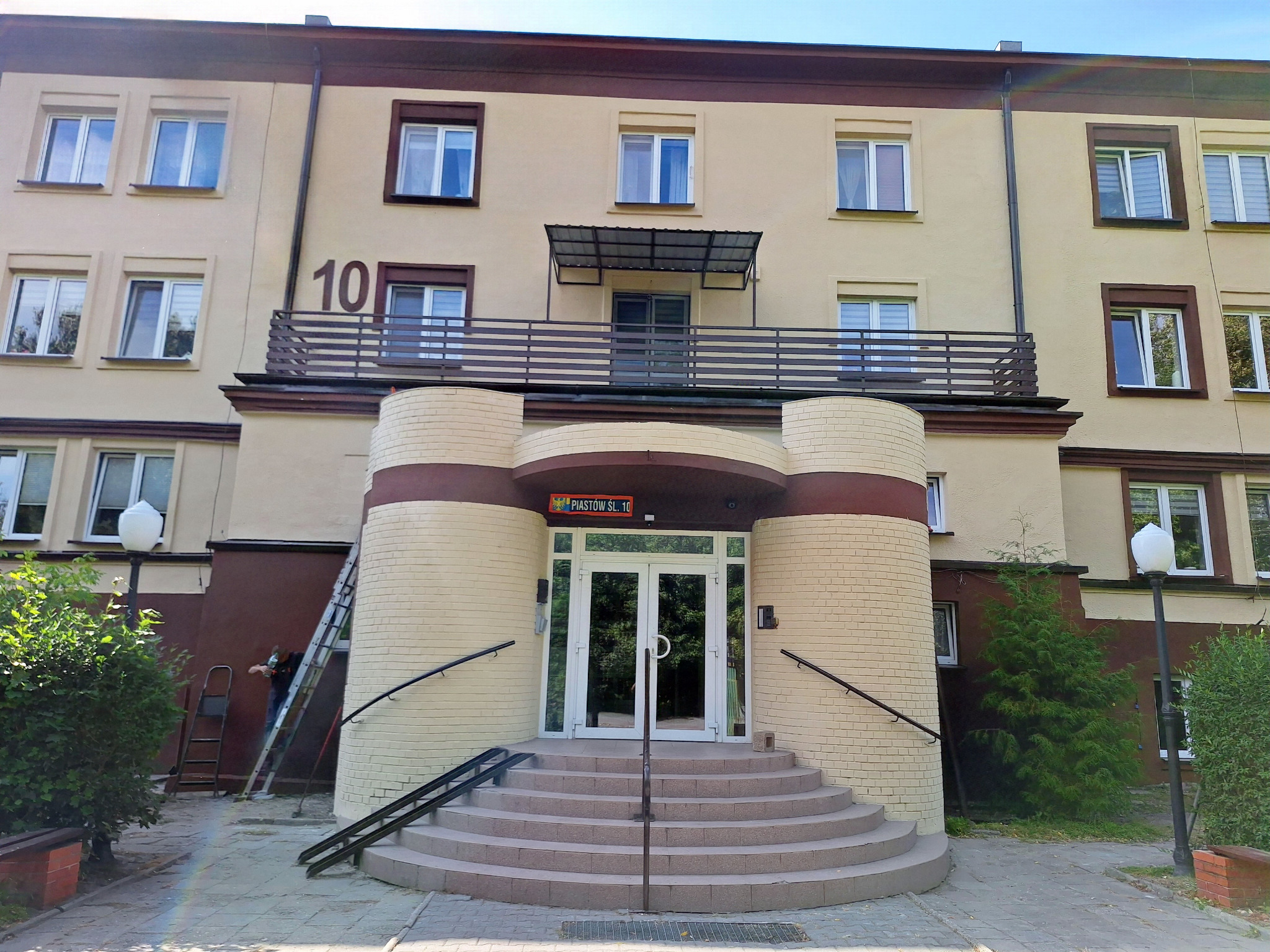 Wejście do budynku z balustradą i poręczami, widoczne schody i pracownik na drabinie. Balustrada z numerem 10. Elementy metalowe.