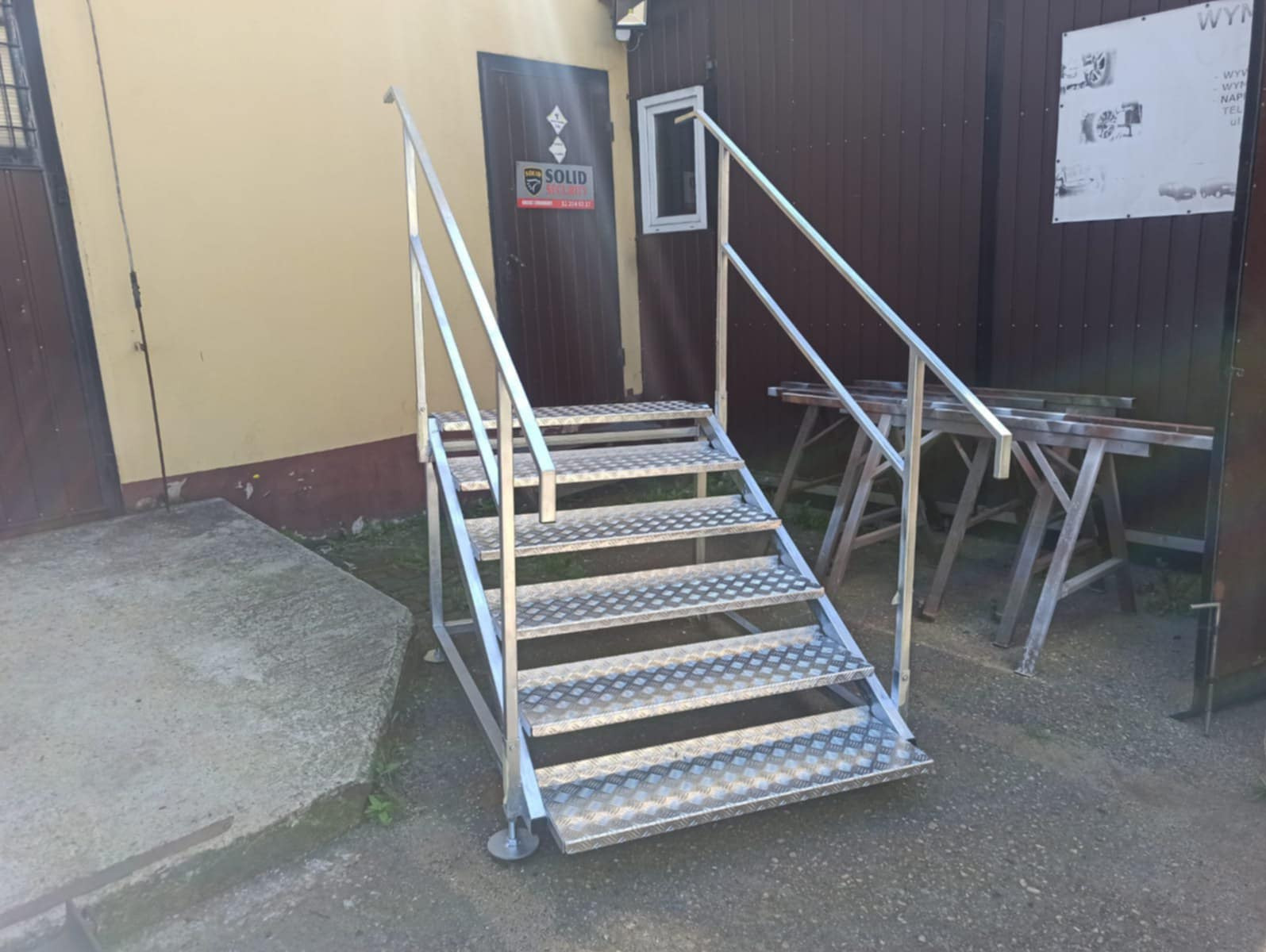 Zewnętrzne schody metalowe z poręczami, wykonane z blachy ryflowanej, prowadzące do drzwi. Konstrukcja spawana, ocynkowana, z regulowanymi stopkami.