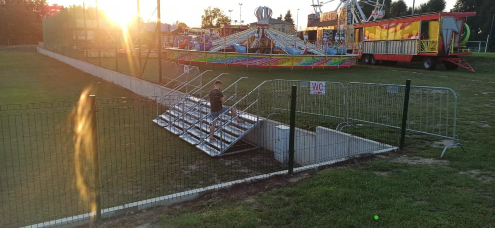 Metalowe schody z poręczami prowadzące do podziemnego pomieszczenia na tle wesołego miasteczka w promieniach zachodzącego słońca.