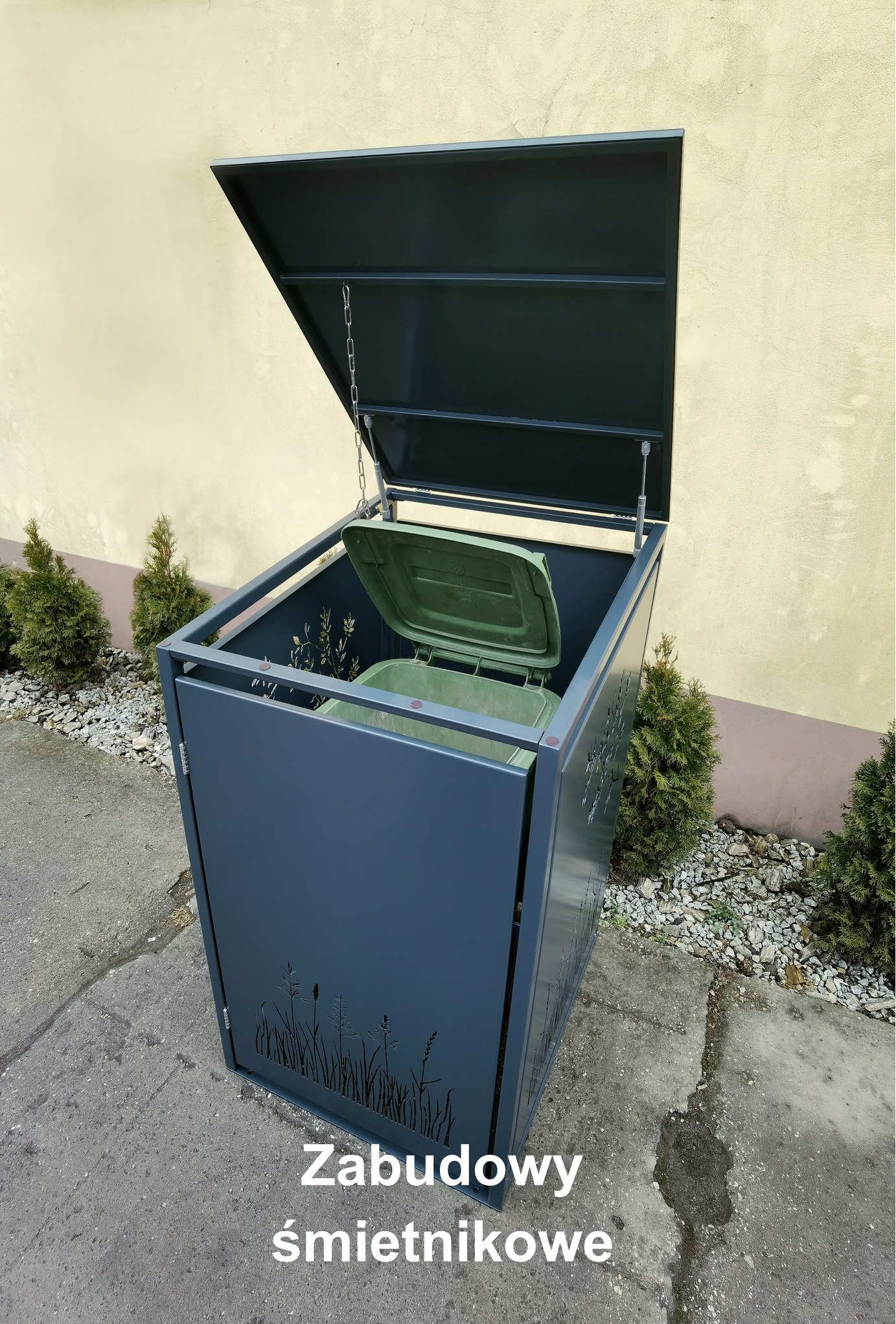 Nowoczesna, grafitowa obudowa na pojemniki na śmieci z motywem roślinnym, z otwartą klapą i widocznym wkładem, ustawiona na zewnątrz budynku.