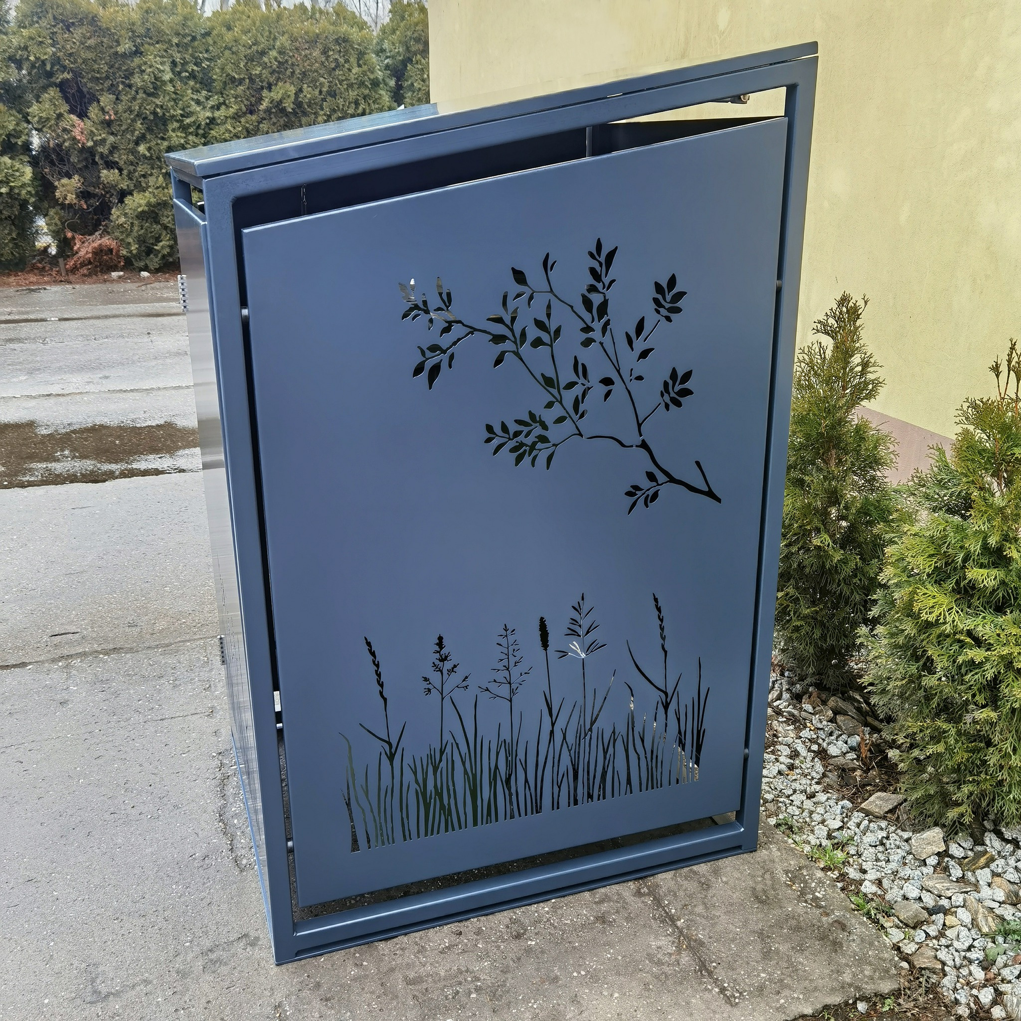 Metalowa obudowa na kosz z dekoracyjnym wzorem roślinnym, ustawiona na betonowym podłożu obok zieleni. Nowoczesny design z precyzyjnie wyciętymi detalami.