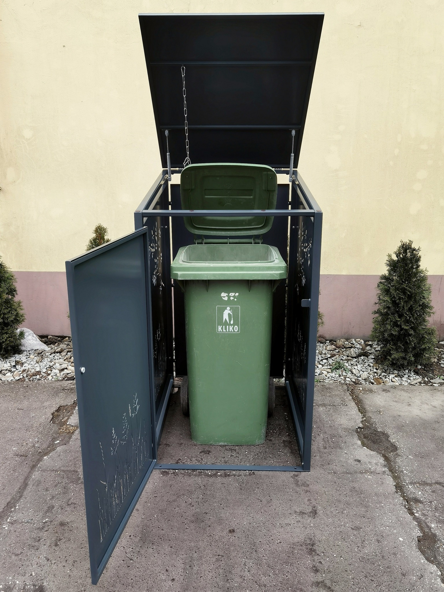 Stalowa obudowa na pojemnik na odpady z dekoracyjnym motywem roślinnym, otwartymi drzwiczkami i podnoszoną pokrywą na łańcuchu, na tle budynku i zieleni.
