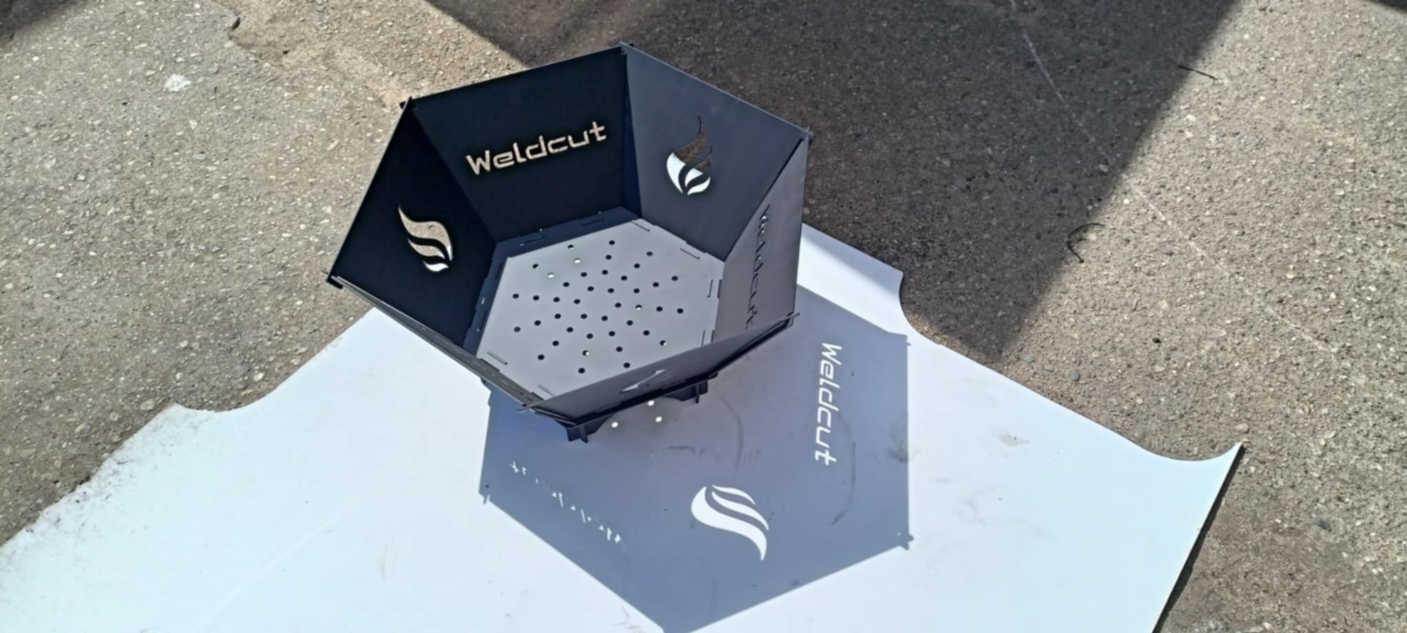 Metalowy kosz na ognisko z wyciętym laserowo logo i wzorem płomienia, ustawiony na białym arkuszu na zewnątrz. Widoczne perforowane dno i cień.