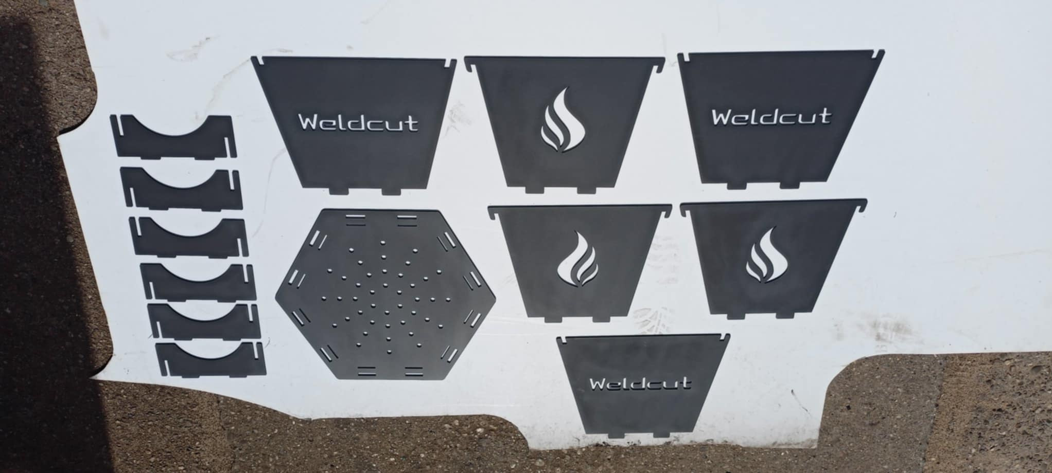 Części wycięte z metalu na palniku CNC, gotowe do spawania. Elementy z logo i motywem płomienia, leżą na białym tle. Widoczny fragment podłoża.