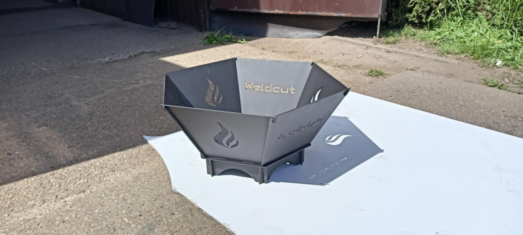 Metalowy kosz na ognisko z wyciętym logo 'Weldcut' i motywem płomieni, ustawiony na białej powierzchni na zewnątrz.