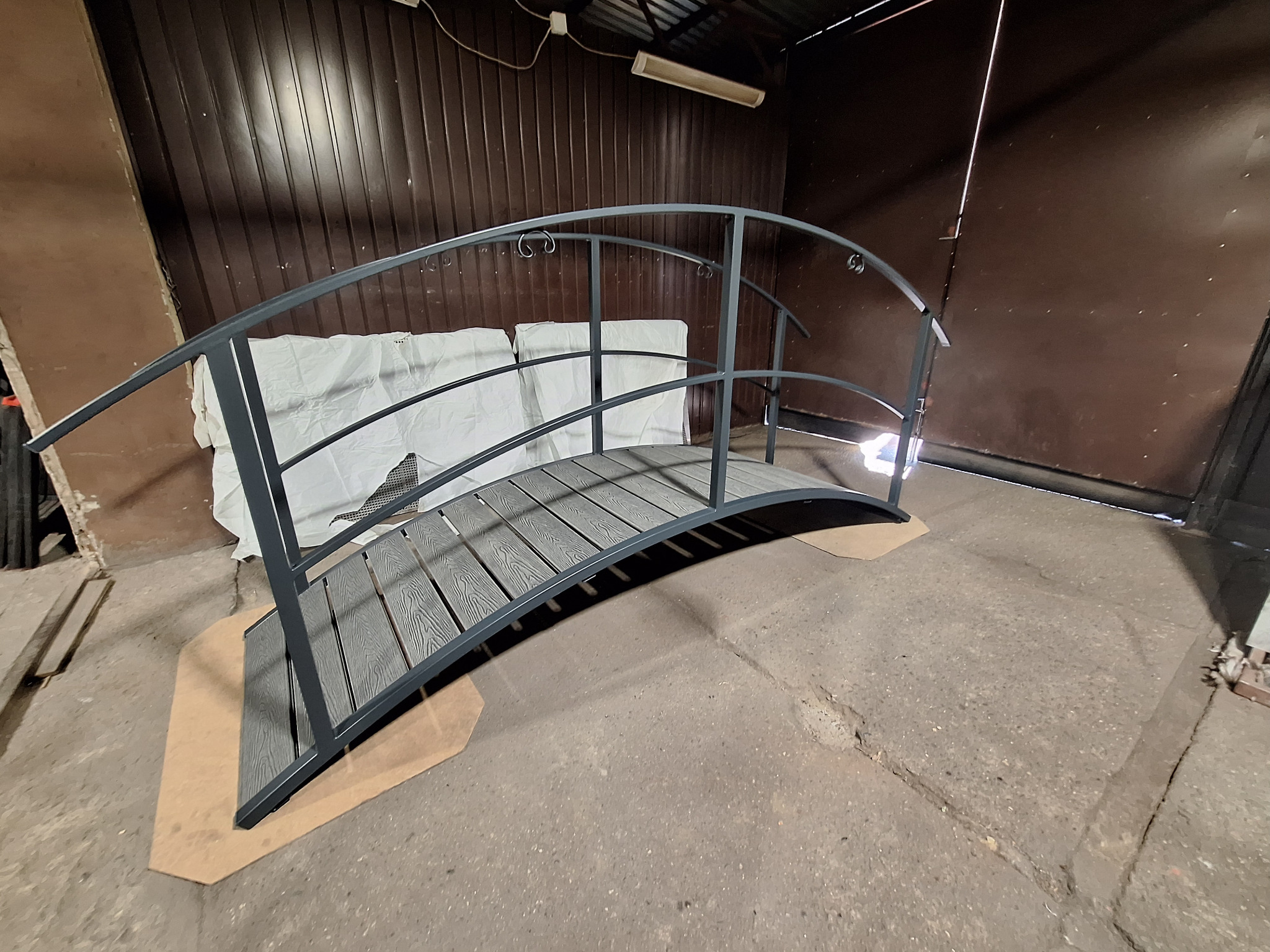 Metalowy mostek ogrodowy z szarą, drewnopodobną podłogą, widoczny w warsztacie. Konstrukcja łukowa z poprzecznymi elementami pełni funkcję dekoracyjną.