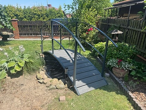 Metalowy mostek z poręczami nad oczkiem wodnym w ogrodzie, otoczony zielenią i kamieniami. Całość wkomponowana w krajobraz.