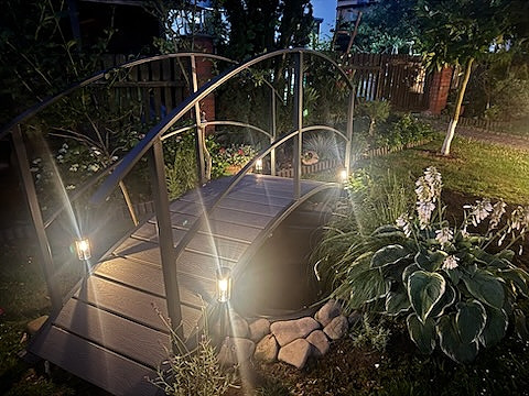 Oświetlony mostek z metalową konstrukcją w ogrodzie, z dekoracyjnymi lampkami i kamieniami. Ujęcie nocne, podkreślające nastrojowe oświetlenie.