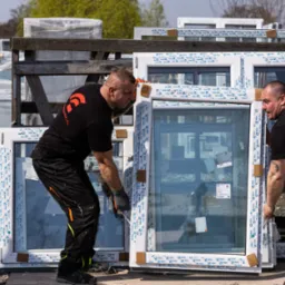 Dw&oacute;ch pracownik&oacute;w w czarnych koszulkach z logo firmy przenosi białe okna PCV, zabezpieczone pianką i folią, z samochodu dostawczego na zewnątrz.