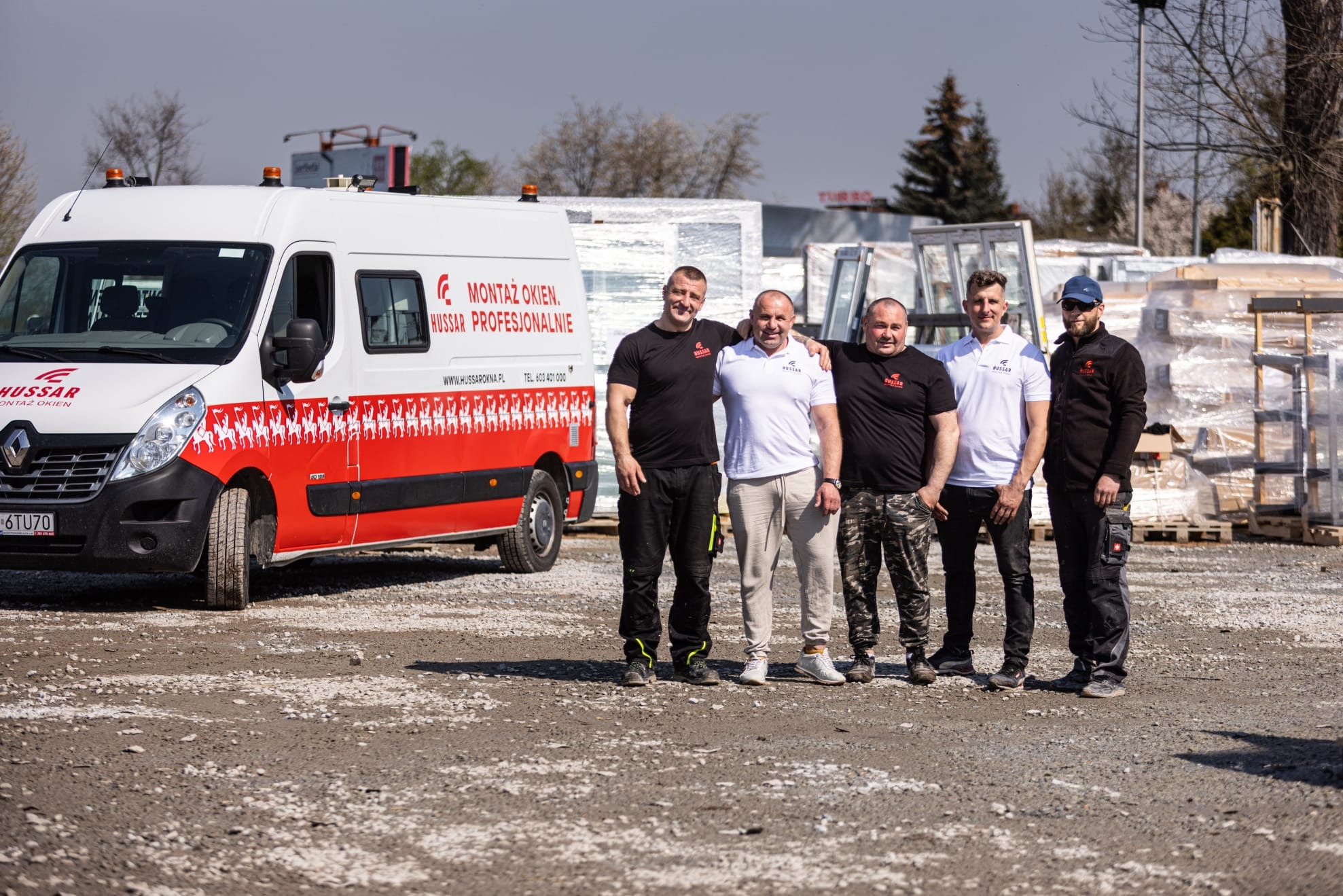 Ekipa montażowa Hussar pozuje przed samochodem firmowym na tle składowanych okien i bram.
