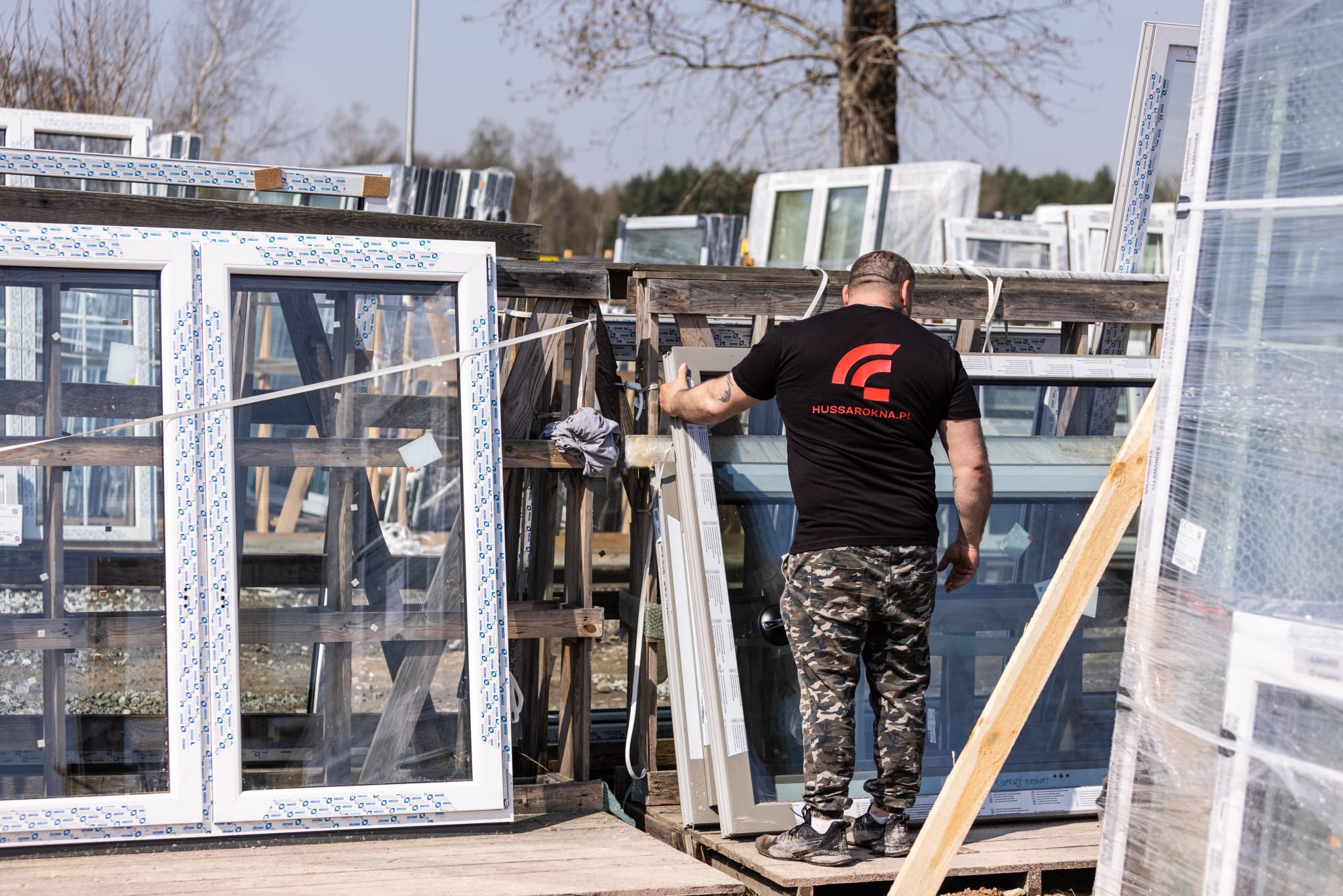 Pracownik w czarnej koszulce z logo firmy HUSSAROKNA.pl przenosi okno PCV na drewnianym regale, w tle inne okna, niektóre w folii ochronnej.