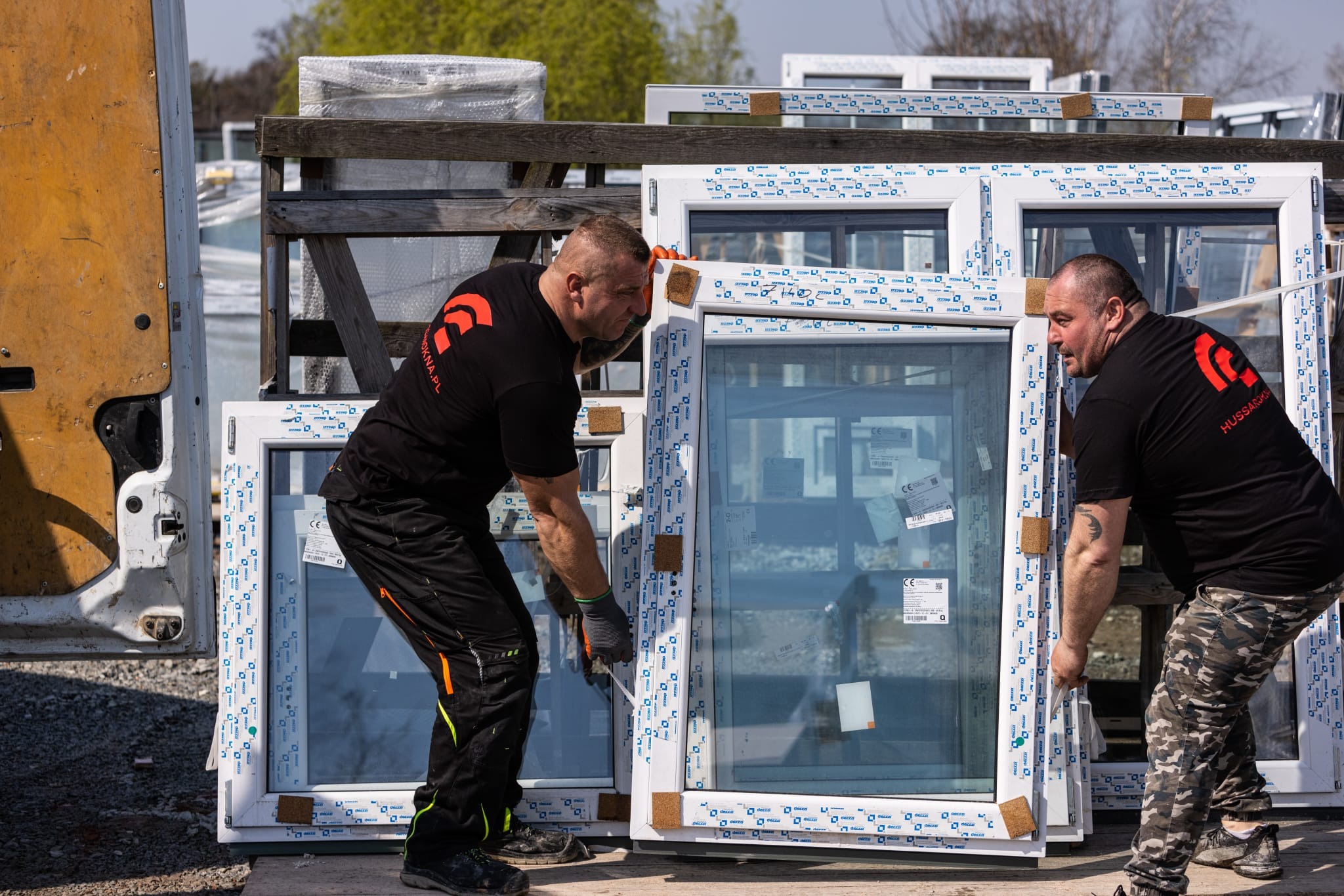 Dwóch pracowników w czarnych koszulkach z logo firmy przenosi białe okna PCV, zabezpieczone pianką i folią, z samochodu dostawczego na zewnątrz.