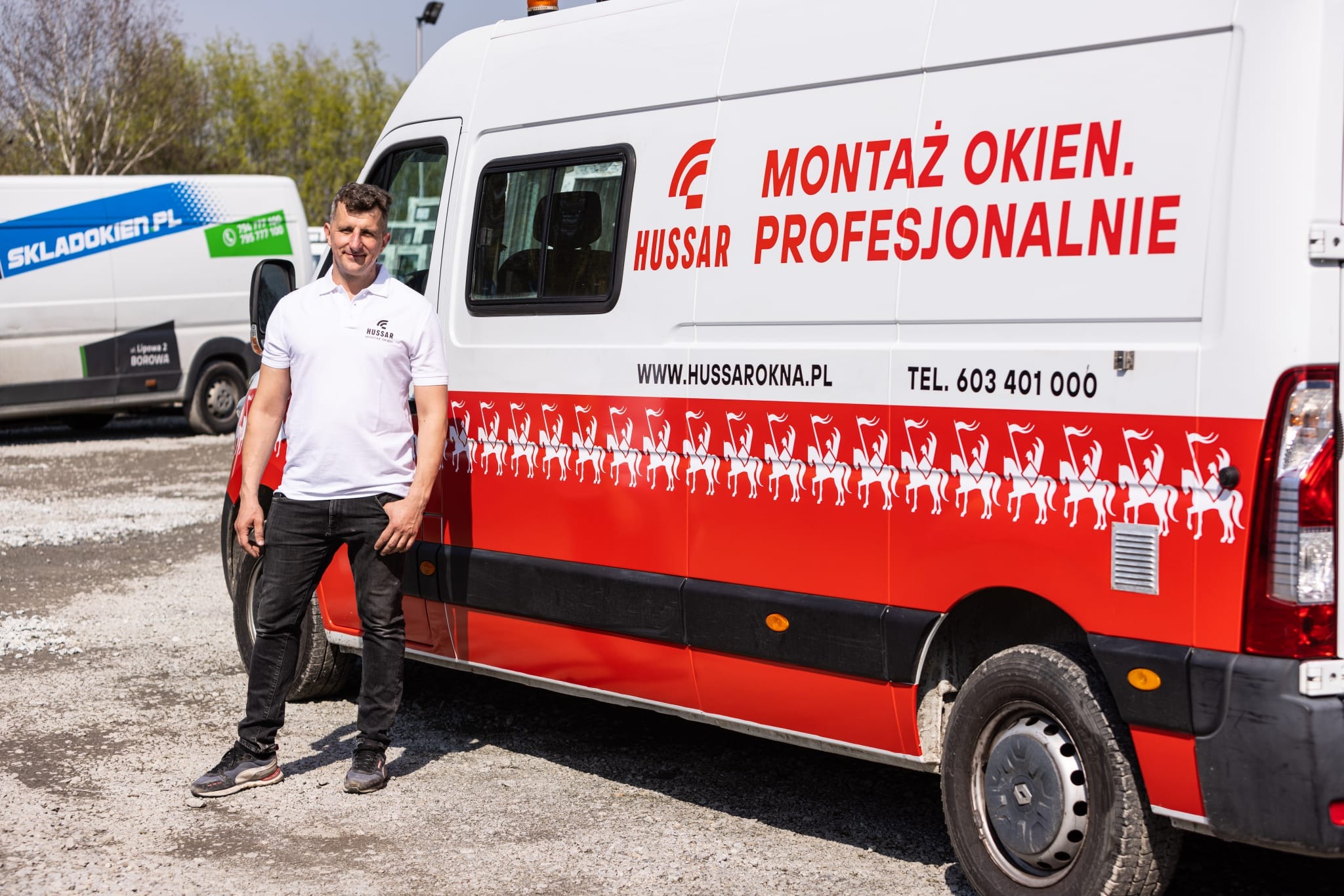 Mężczyzna w białej koszulce polo stoi na żwirowym parkingu obok białego samochodu dostawczego z czerwonym pasem i napisem 'Montaż okien Hussar Profesjonalnie' oraz grafiką husarii.